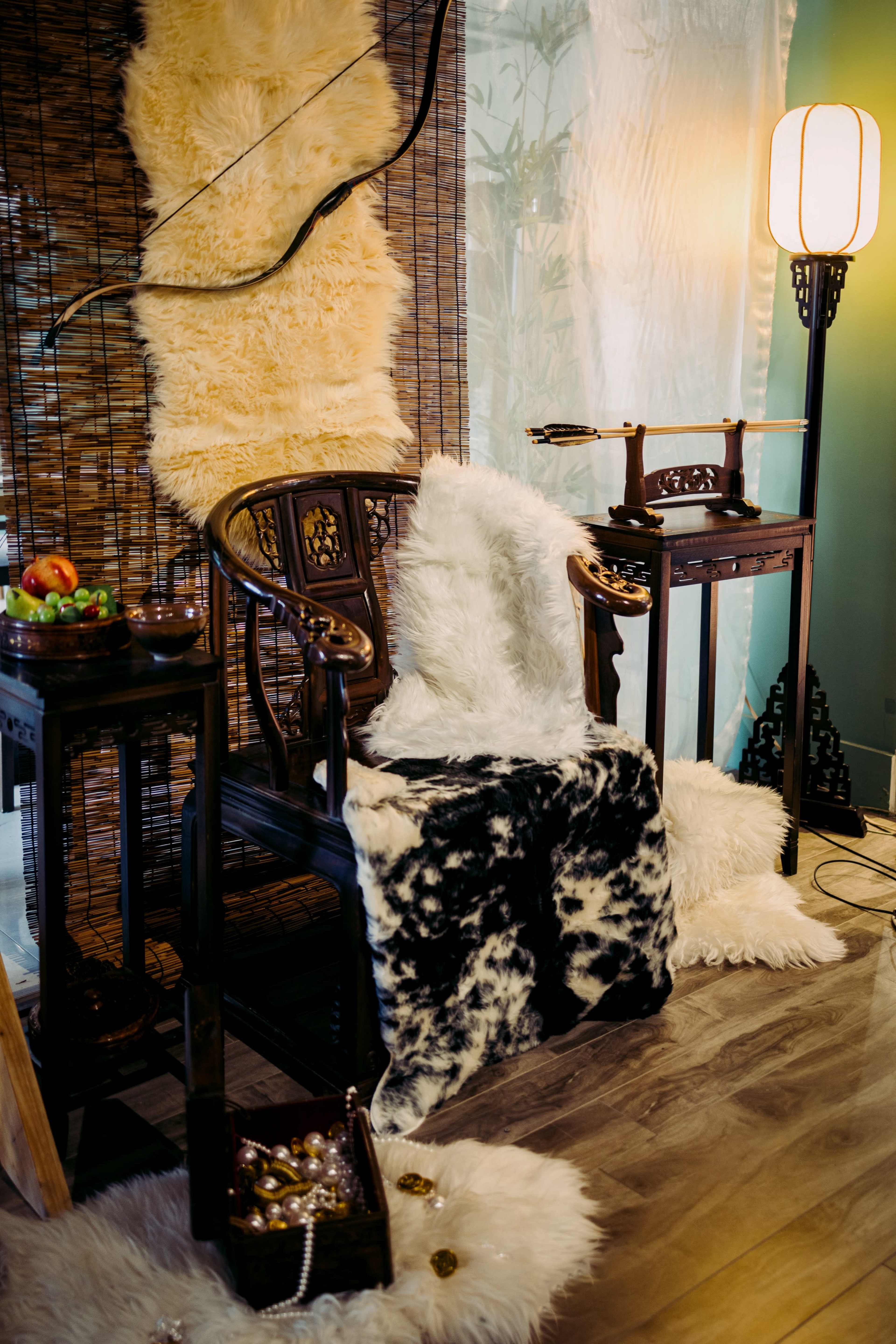 A cozy corner featuring a wooden chair draped with faux fur, accompanied by small wooden tables and decorative items, with a soft light illuminating the space.