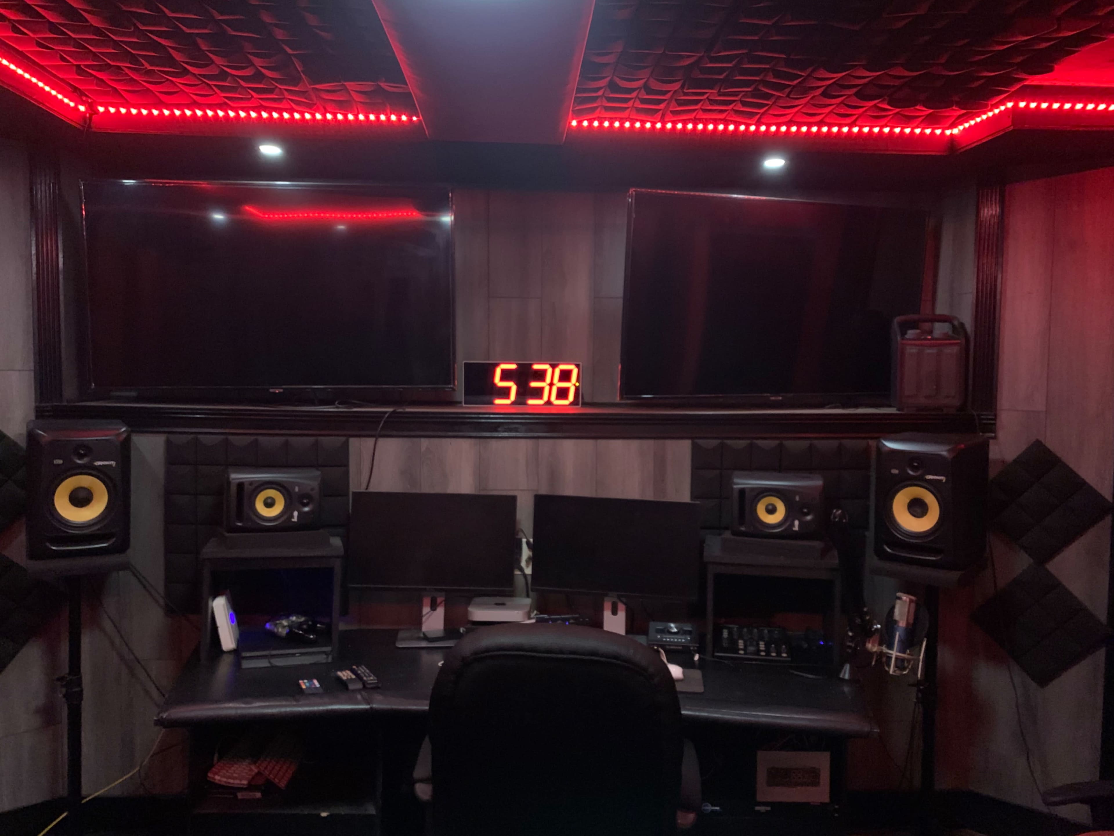 A media production studio features two large screens, a digital clock displaying 5:38, and audio equipment flanking a central workstation.