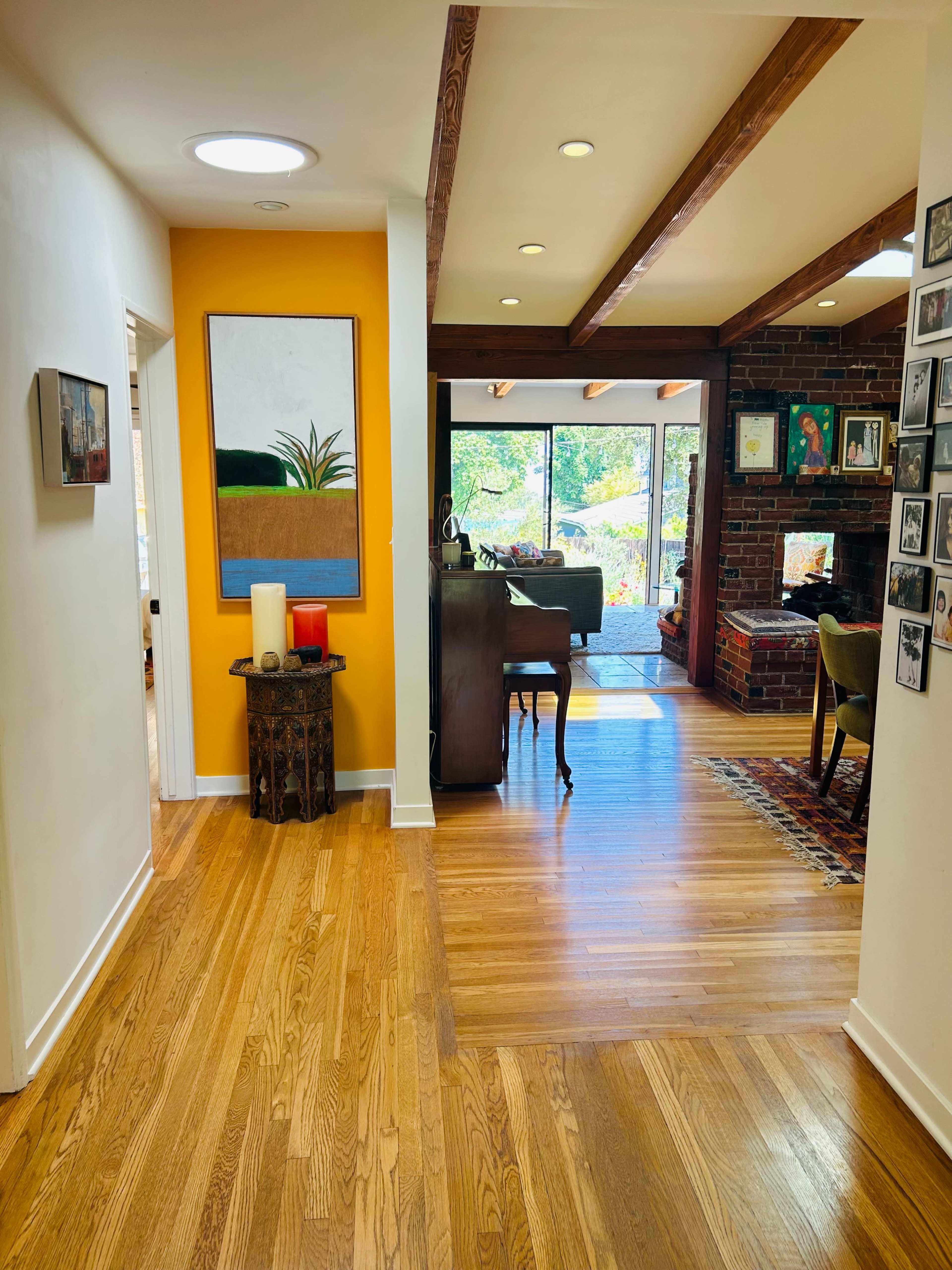 A bright hallway leading to a living area with a view of outdoor greenery, featuring wooden beams and a fireplace.