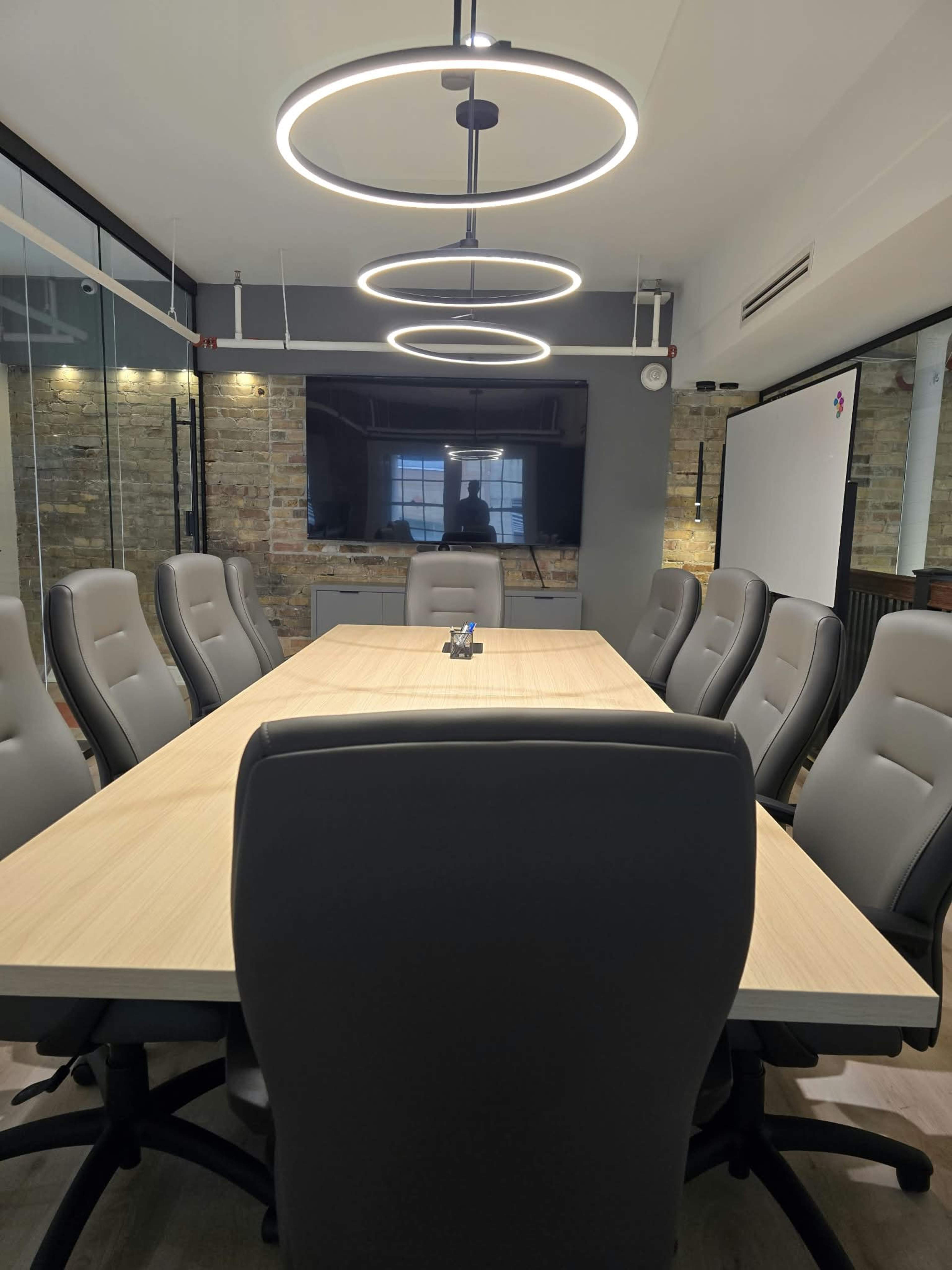 A modern conference room features a long wooden table surrounded by ergonomic chairs and illuminated by circular pendant lights.