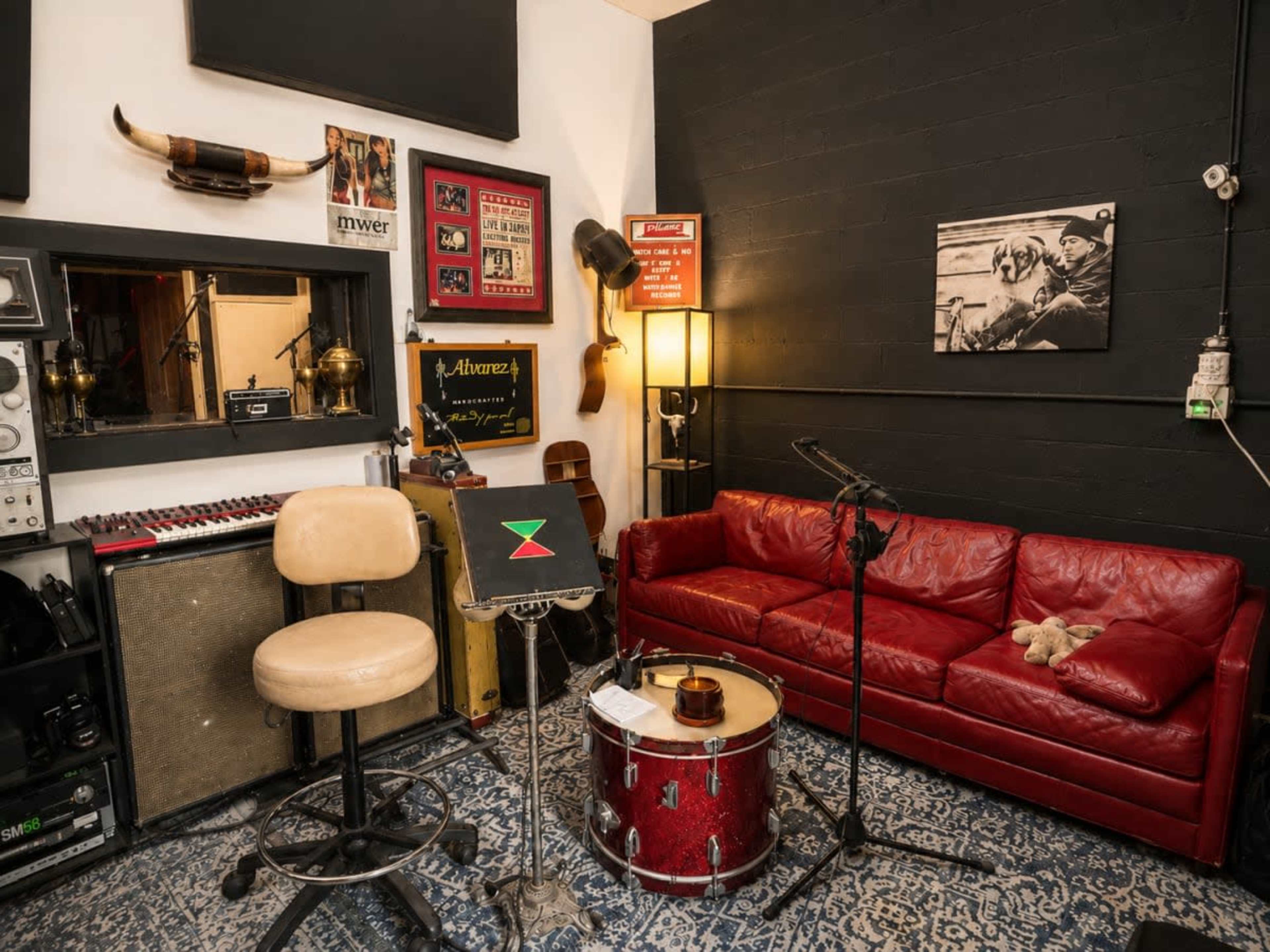 The image shows a small music studio or rehearsal space featuring a red couch, a drum set, a microphone stand, and various musical equipment on display.
