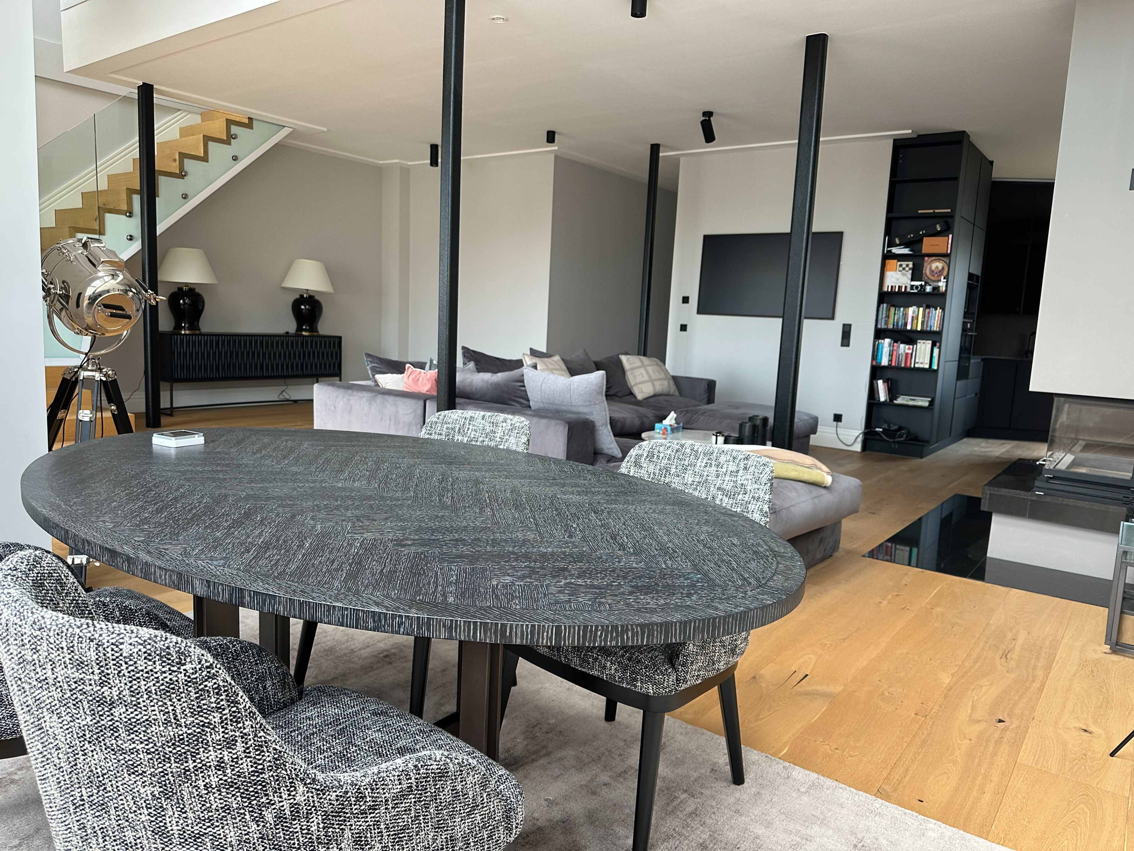 The image shows a modern living space featuring a round dining table with textured chairs, a sectional sofa, and a staircase in the background.