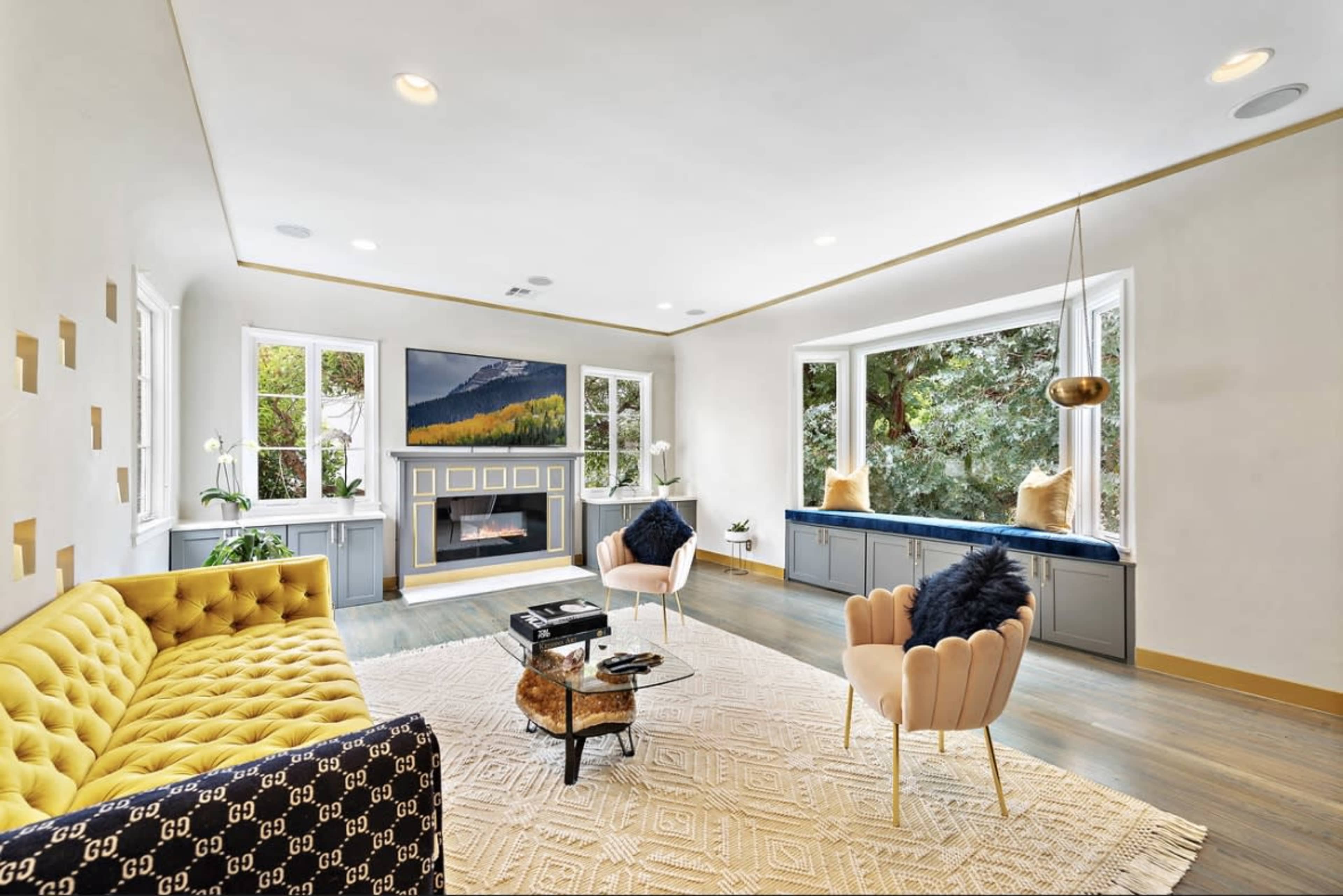 The image depicts a modern living room featuring a yellow tufted sofa, a dark coffee table, and large windows with decorative cushions.