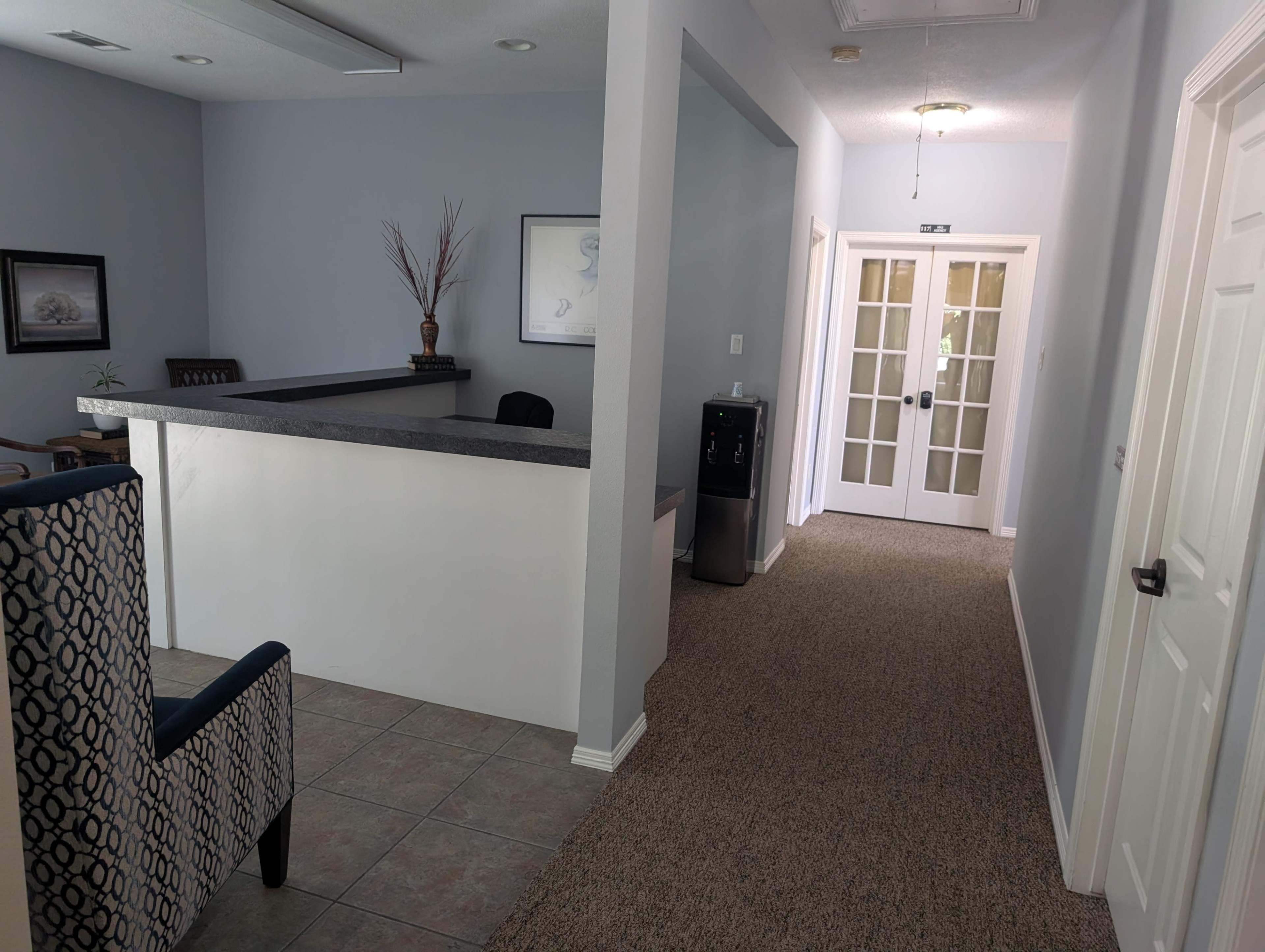 The image shows a reception area with a desk on one side and a hallway leading to double doors on the other, flanked by a water cooler.