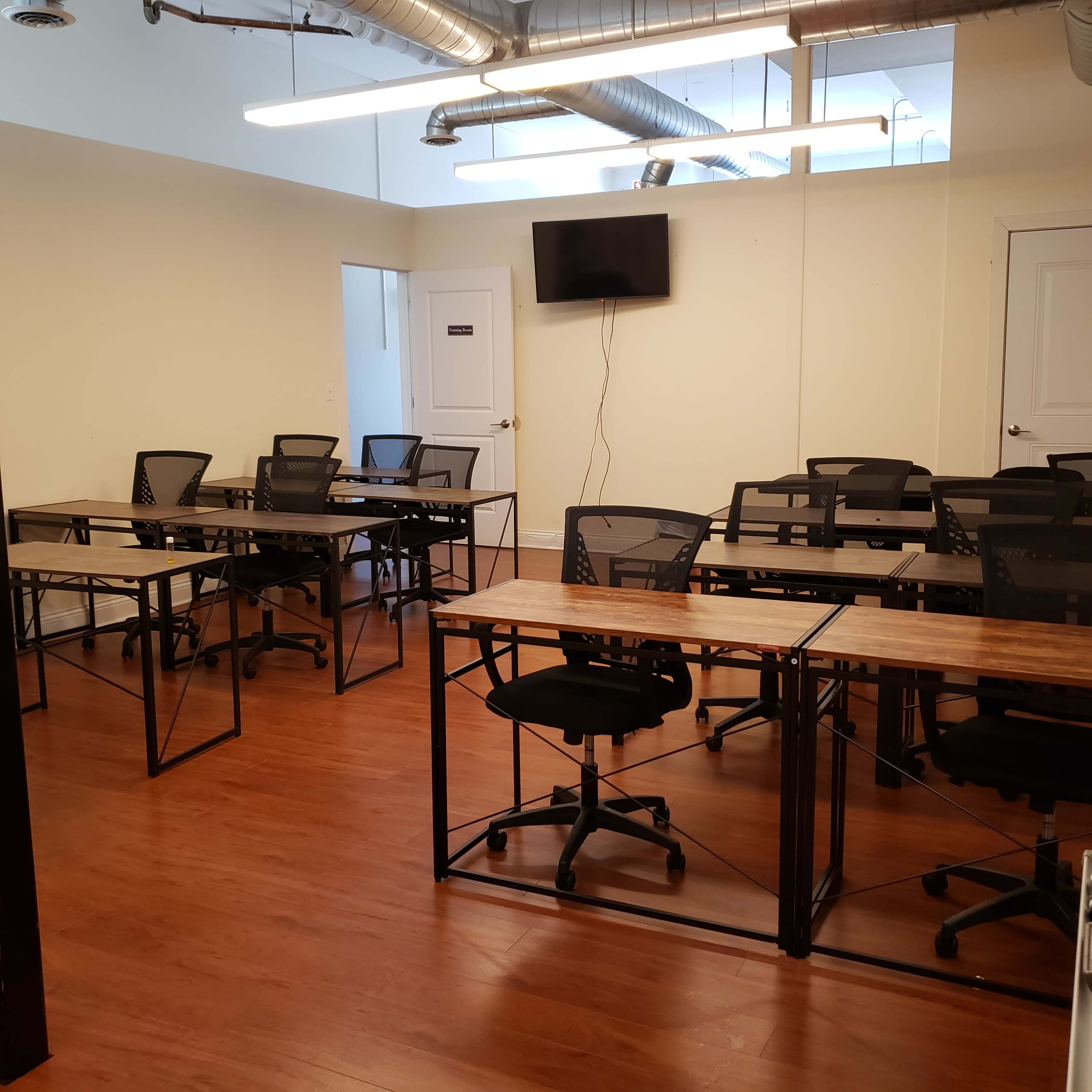 A classroom with several black mesh chairs and desks arranged in a grid layout, and a wall-mounted television in one corner.