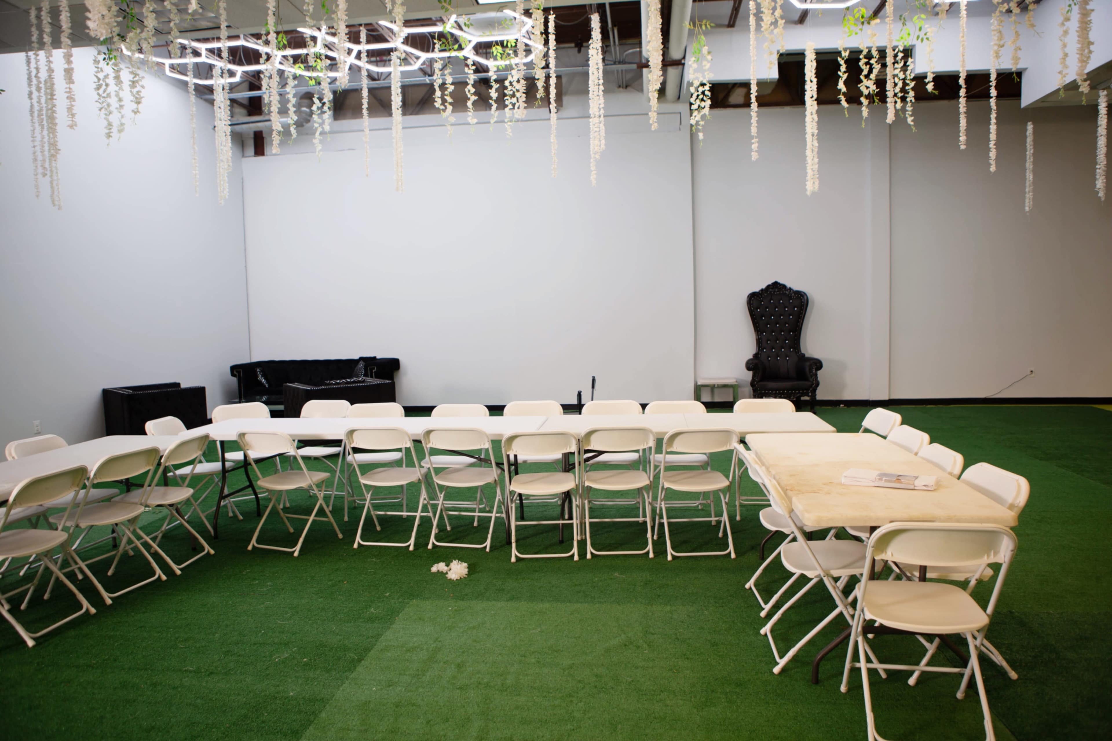 A room with green carpet features a long table surrounded by white folding chairs, with a black couch and an ornate black chair in the background.