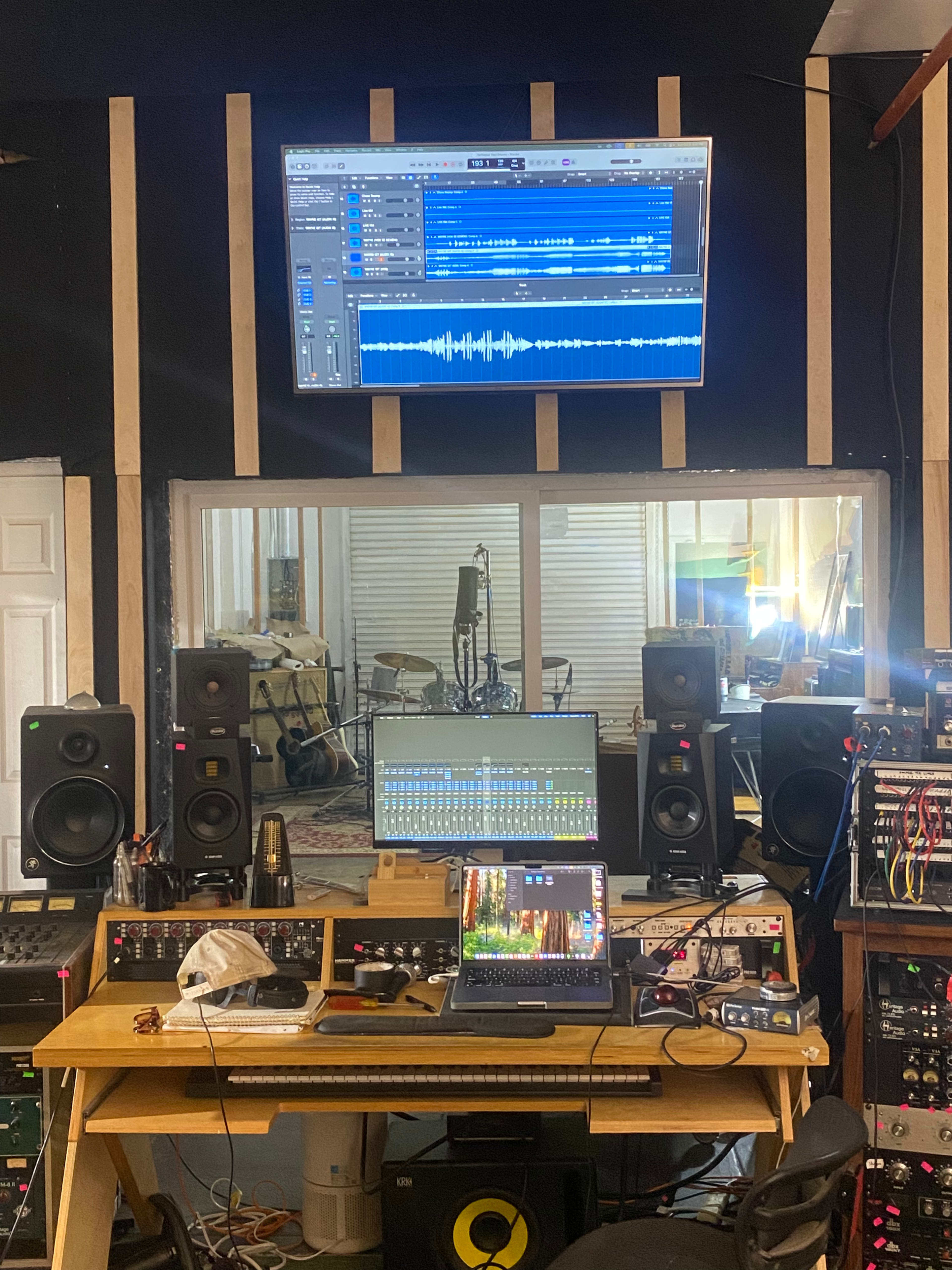 The image shows a music studio workstation with a computer, audio mixing equipment, and studio monitors, all set up in front of a large glass window overlooking a drum kit.