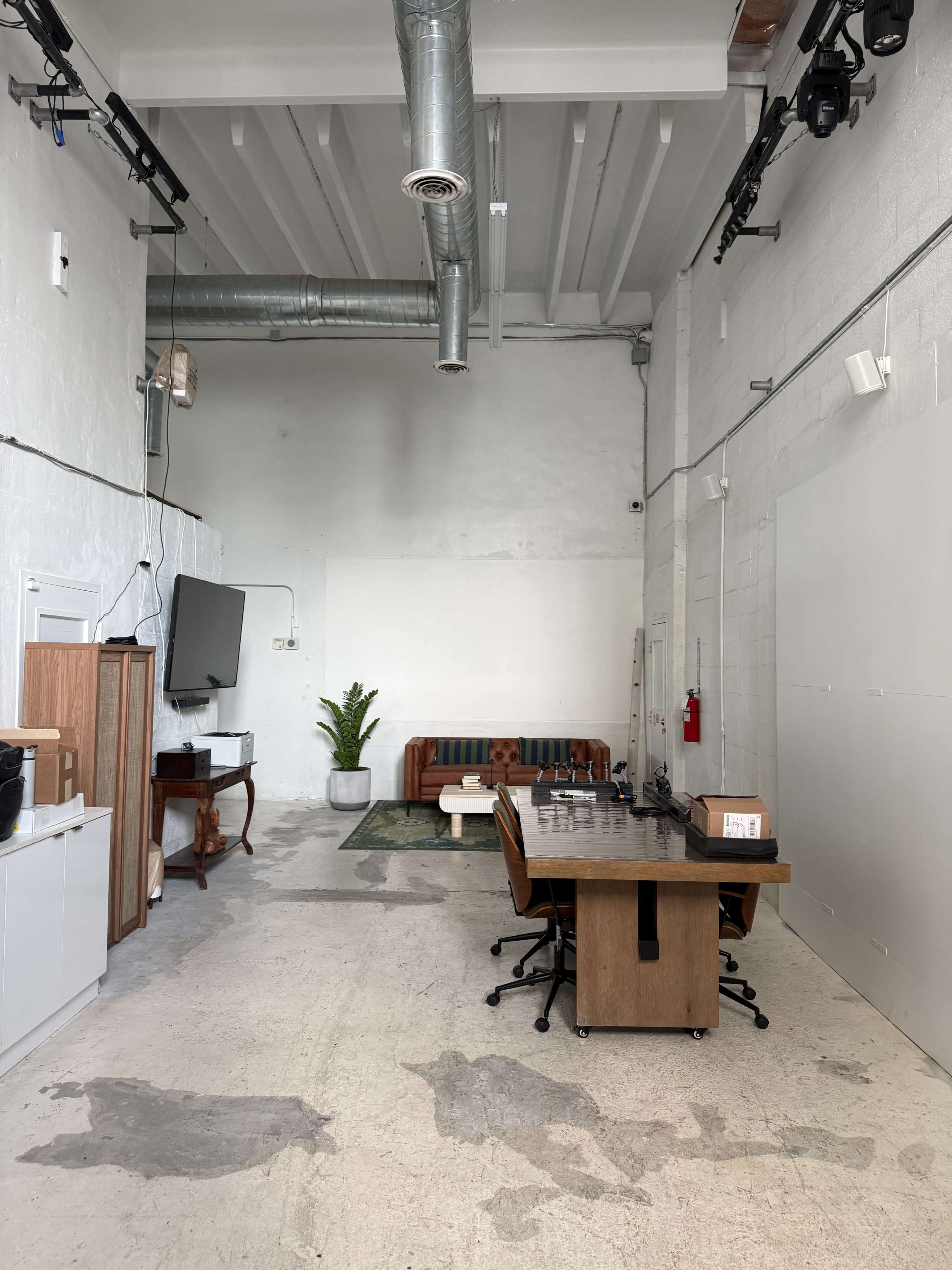 The image shows a spacious, minimalist office interior with a large wooden table, chairs, a sofa, and a potted plant against a light-colored wall.