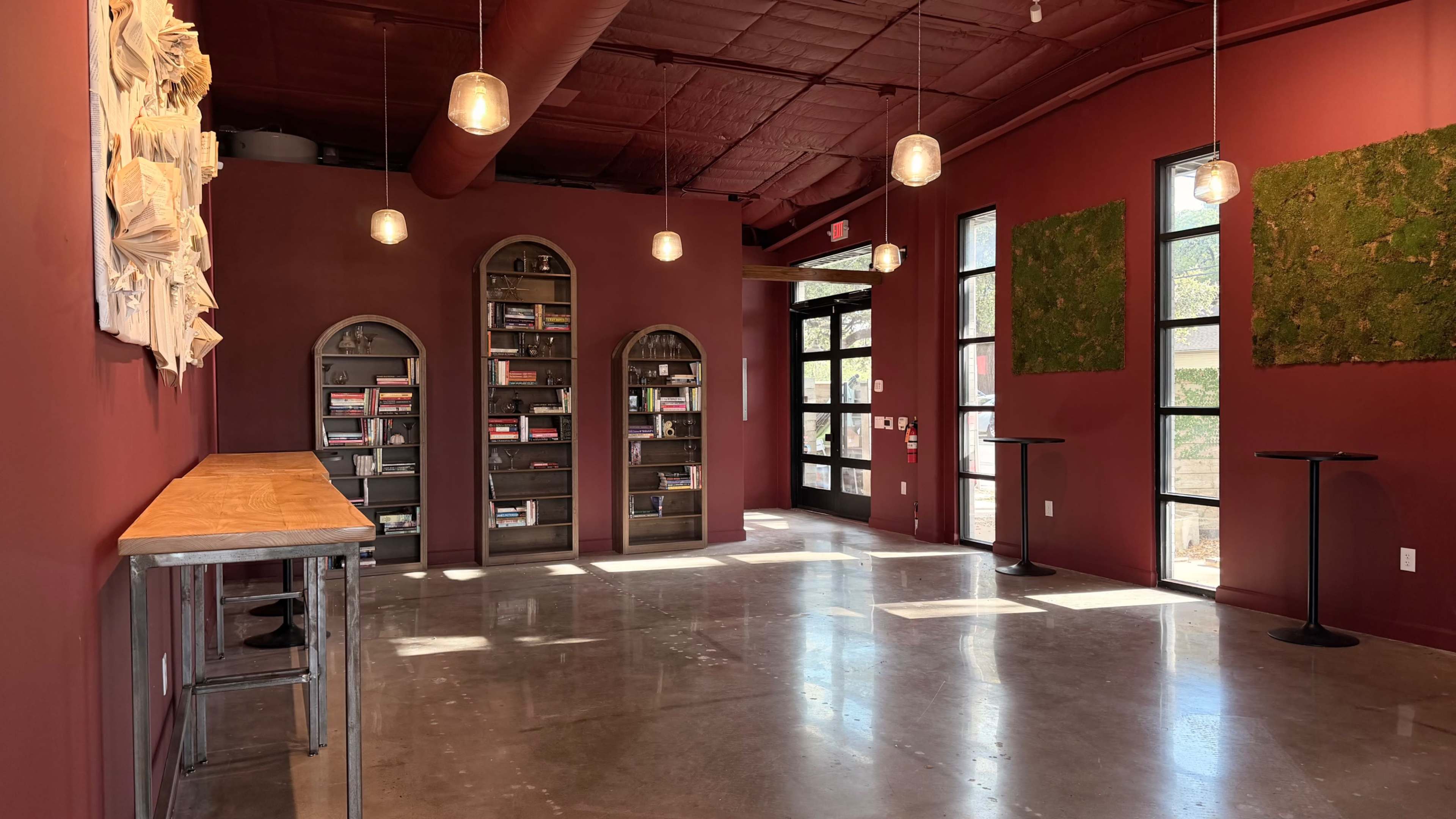 The image shows a modern interior with deep red walls, a wooden counter, two tall tables, a bookshelf filled with books, and illuminated pendant lights.