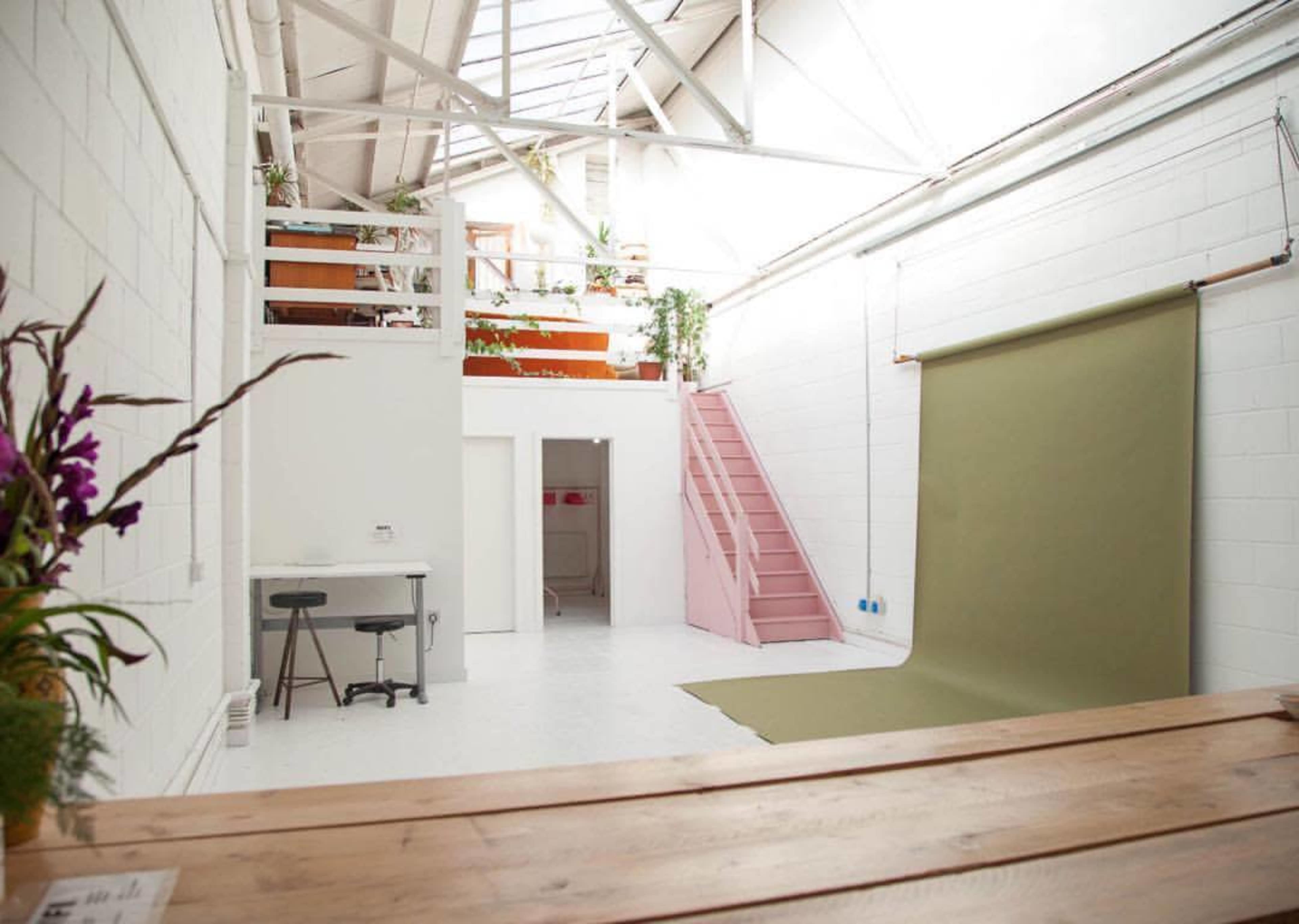 A spacious, well-lit room with white walls, a wooden table, a green backdrop, and pink stairs leading to an upper level adorned with plants.
