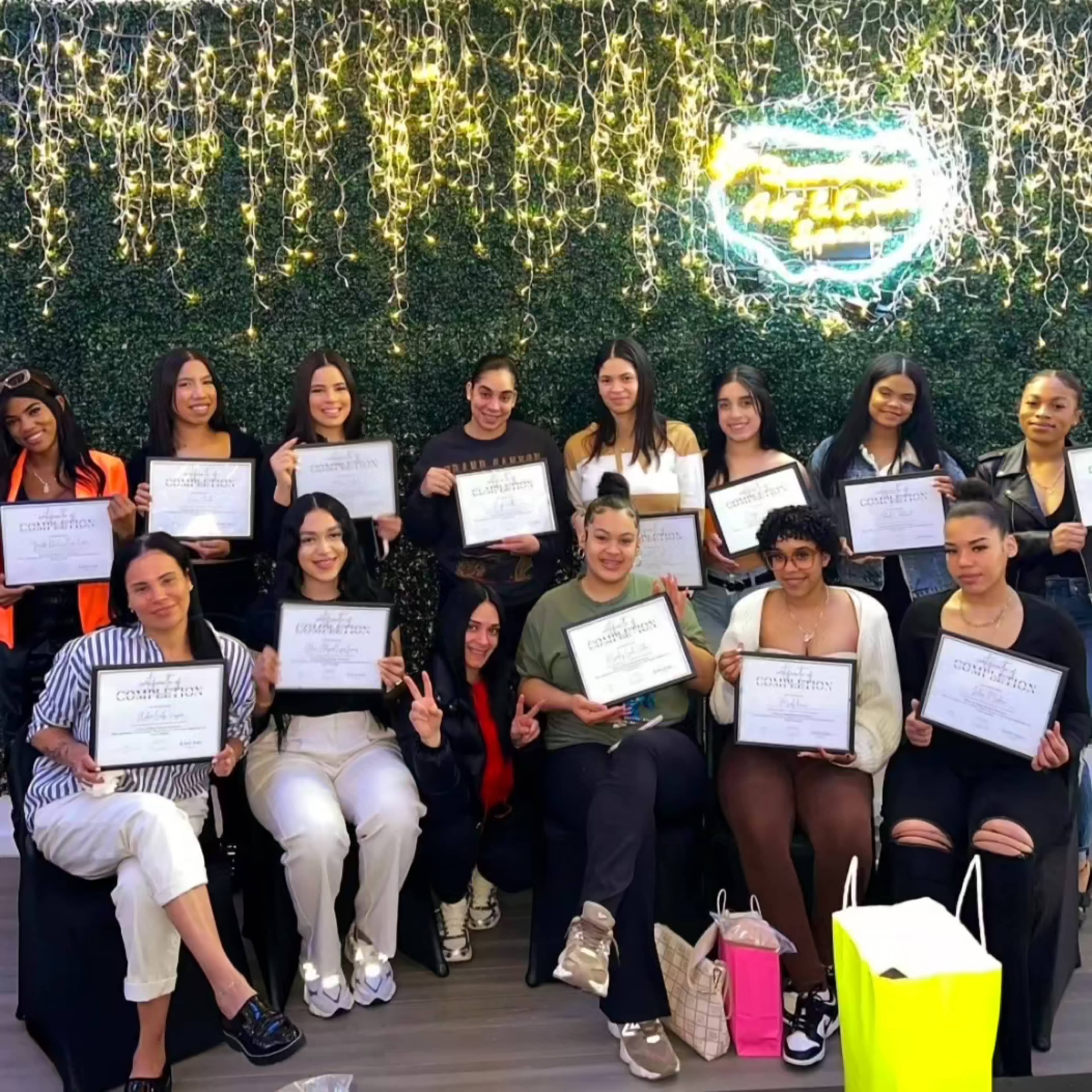 A group of thirteen individuals poses together, each holding a certificate, in front of a backdrop decorated with green foliage and string lights.