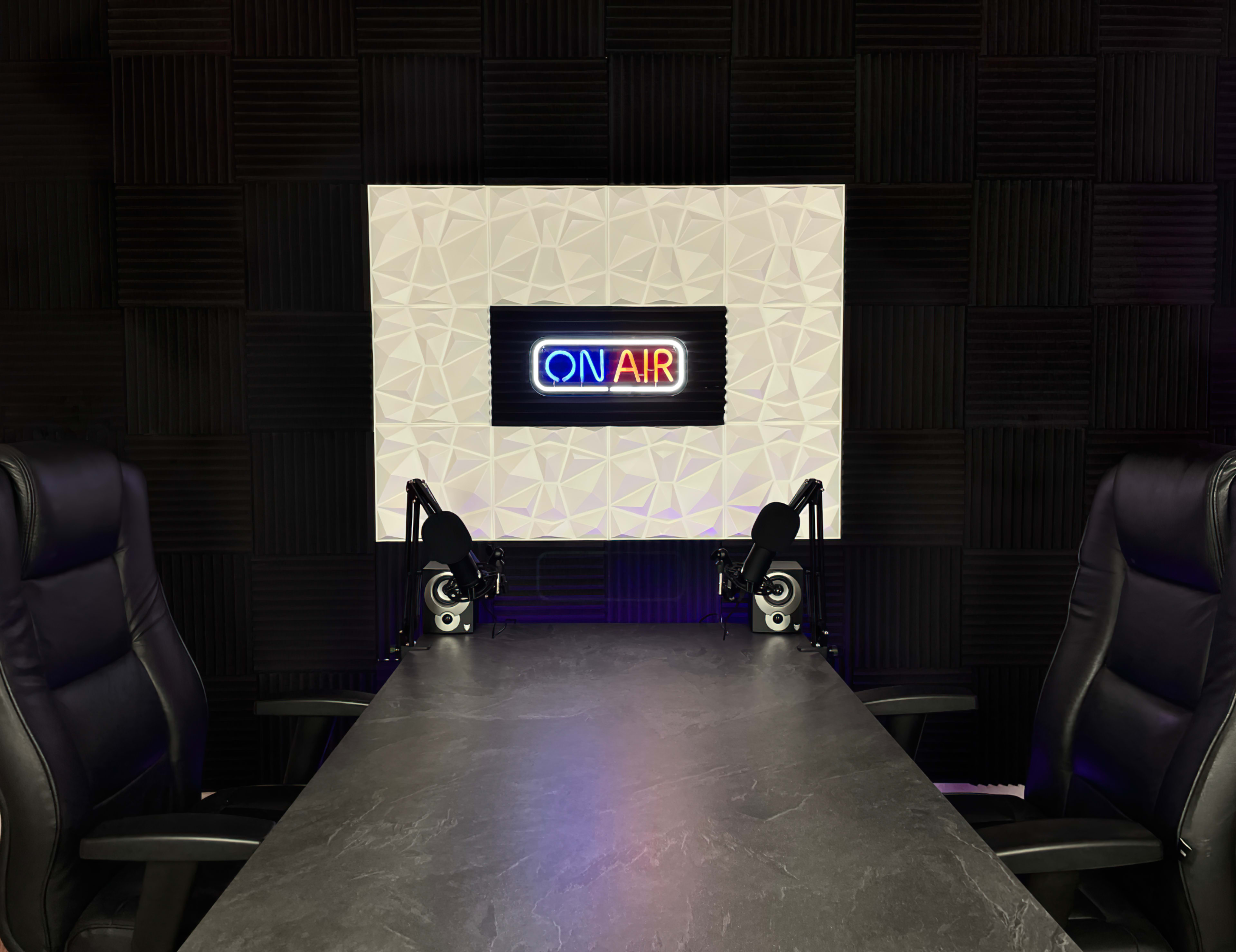 A modern studio setup features a dark table with two chairs facing a illuminated "ON AIR" sign against a textured wall.