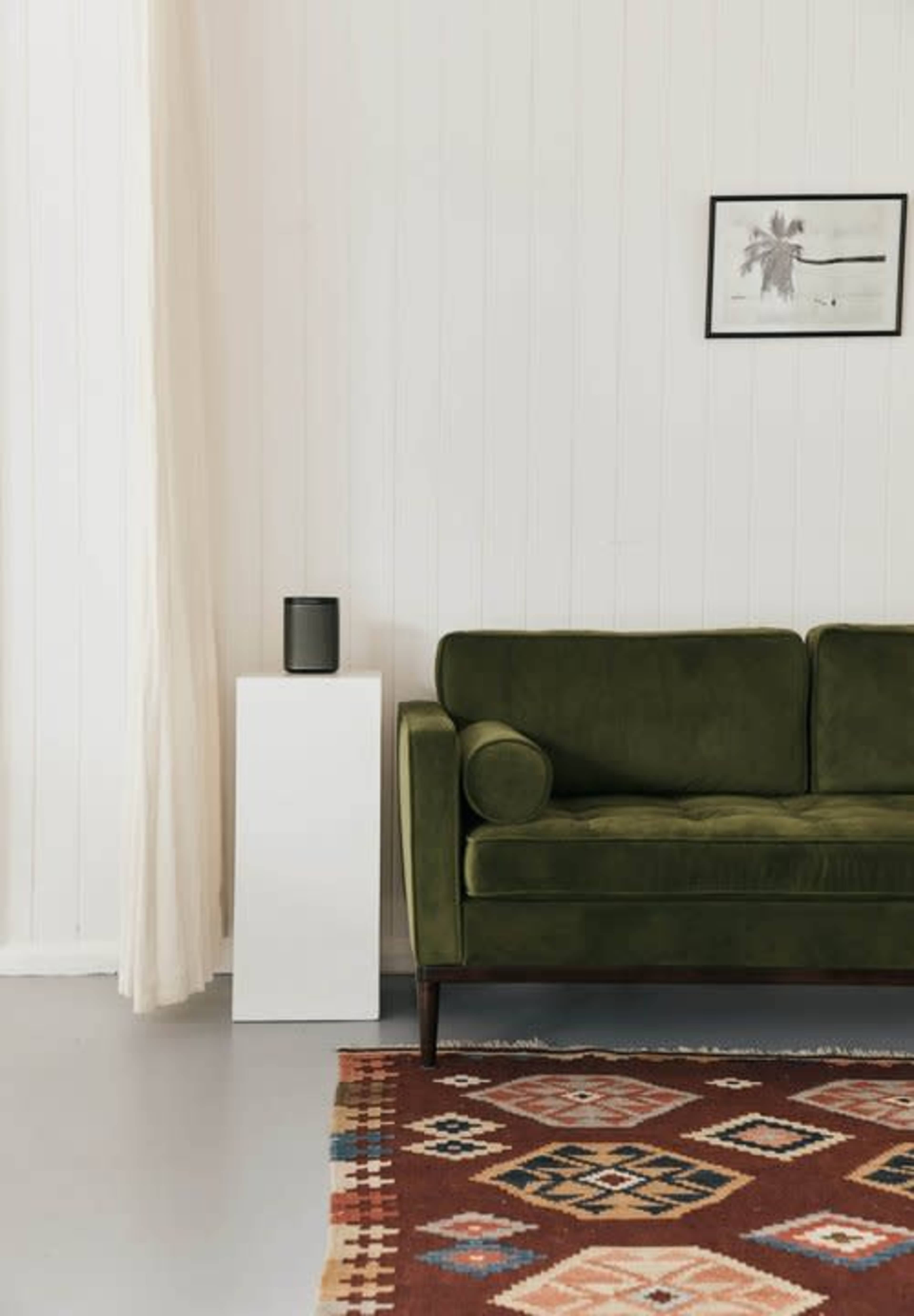 A green velvet sofa sits next to a white side table with a small speaker, on a patterned rug in a bright, minimalistic room.