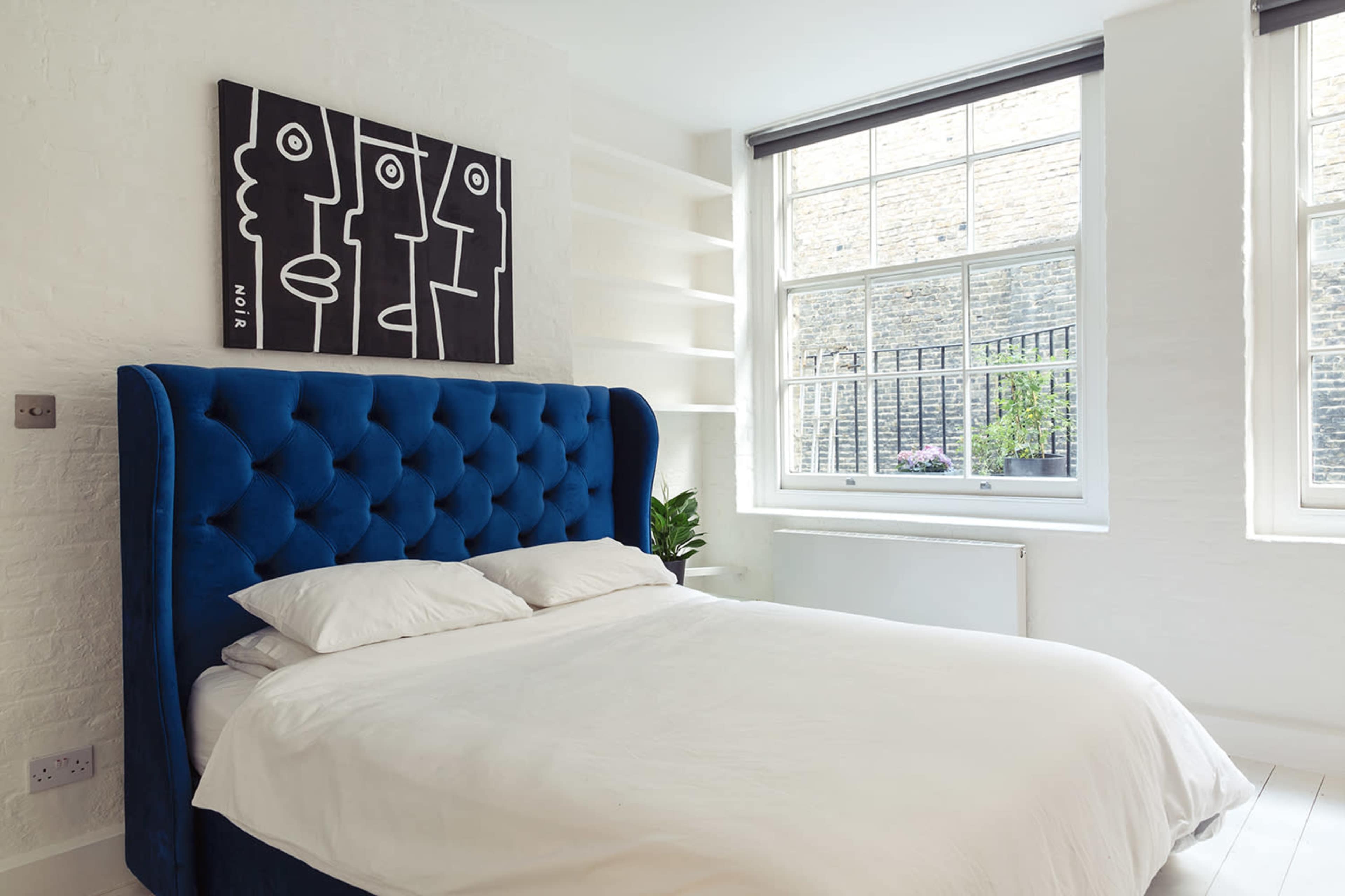 The image shows a bedroom featuring a blue upholstered bed with white bedding, a large window, and a black and white artwork mounted on the wall.