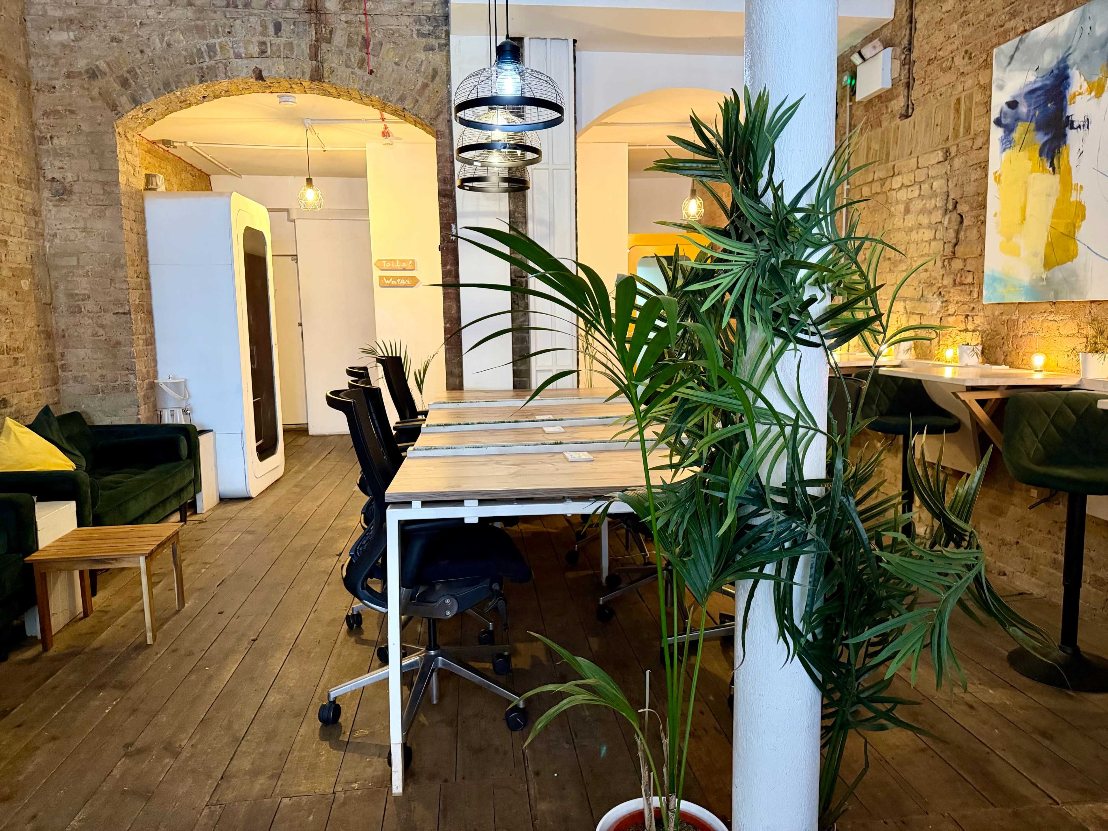 The image shows a spacious coworking area with a large wooden table surrounded by chairs, plants, and a mix of decorative lighting.