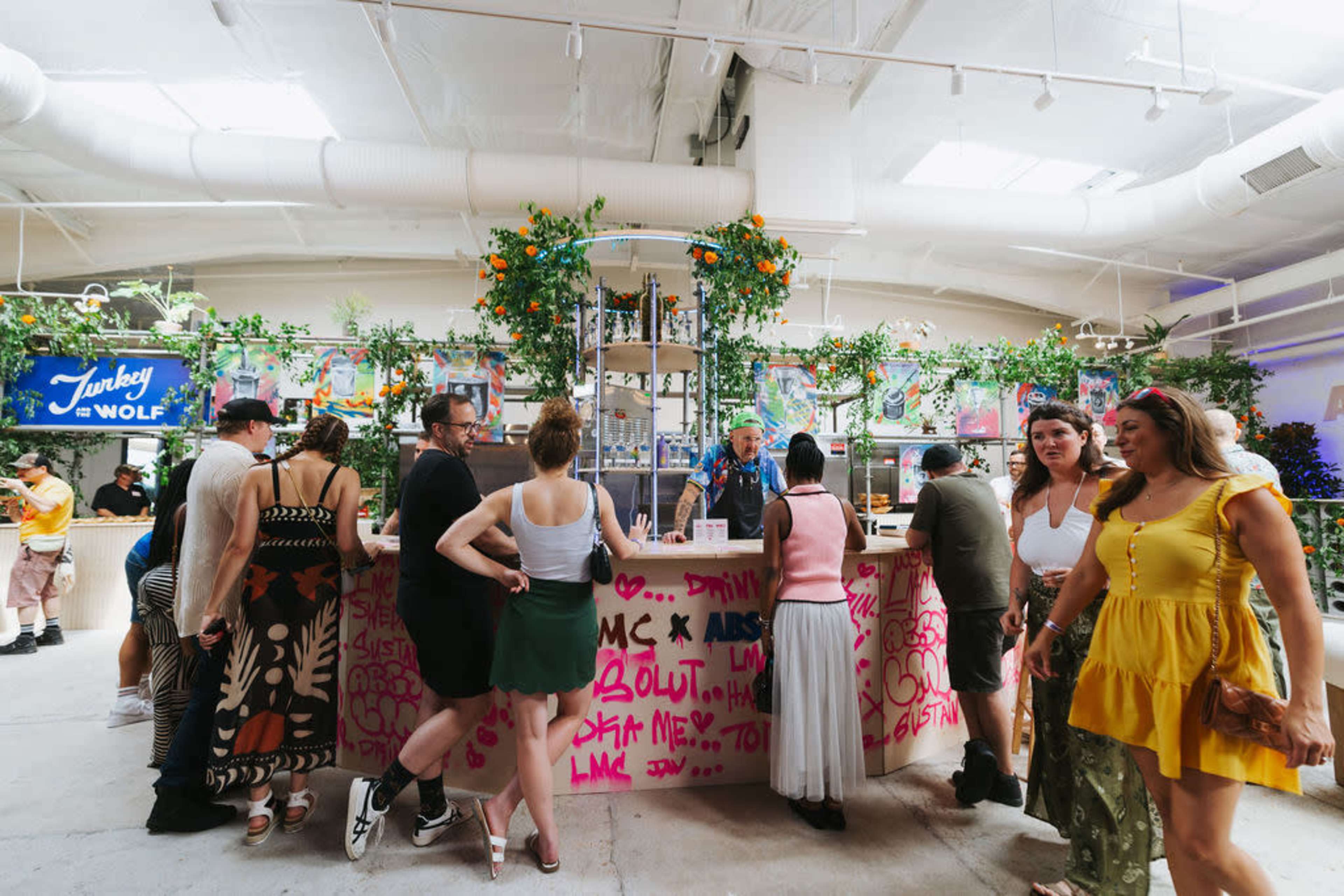 A crowd of people is gathered around a vibrant bar adorned with plants and colorful decorations in a lively indoor setting.
