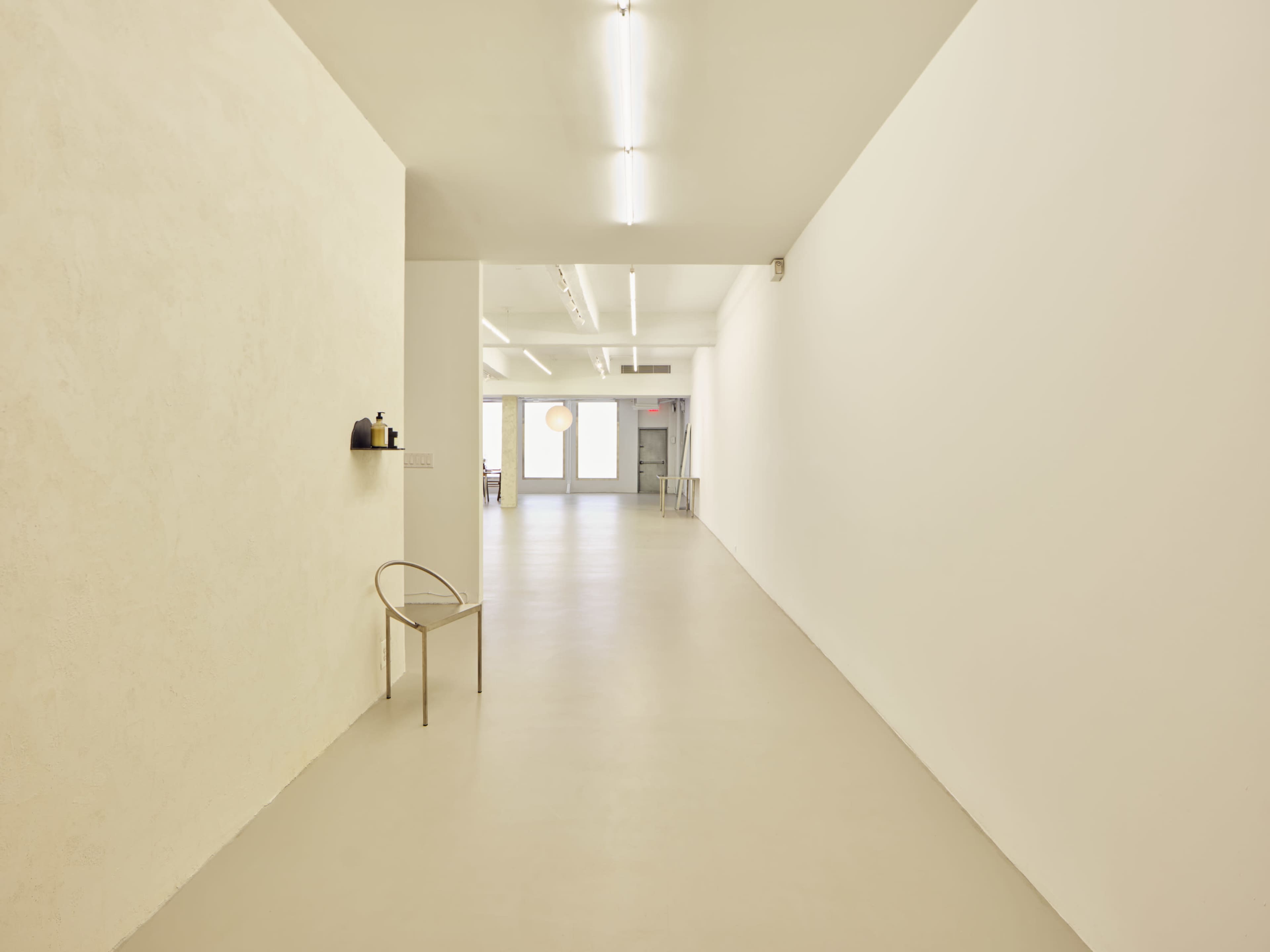 A long, minimalist hallway with white walls and a single chair on the left side.