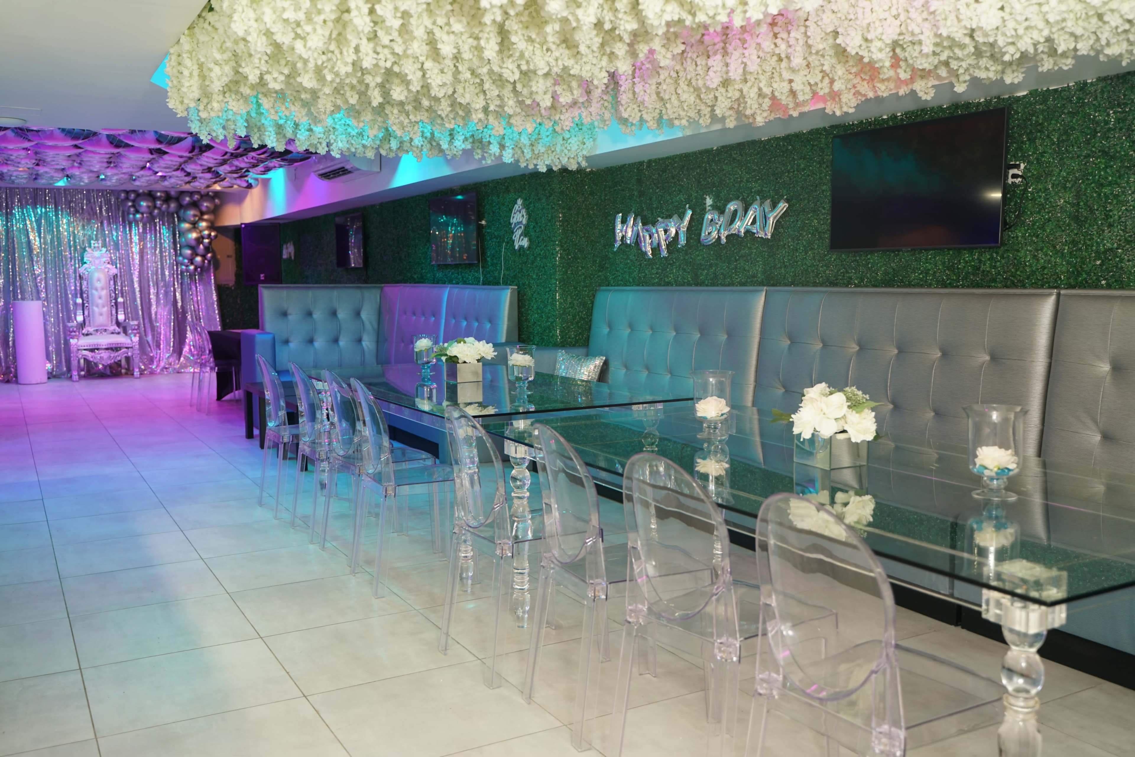 A modern event space with a long glass dining table surrounded by transparent chairs, decorated with white flowers and a backdrop of shimmering fabric.