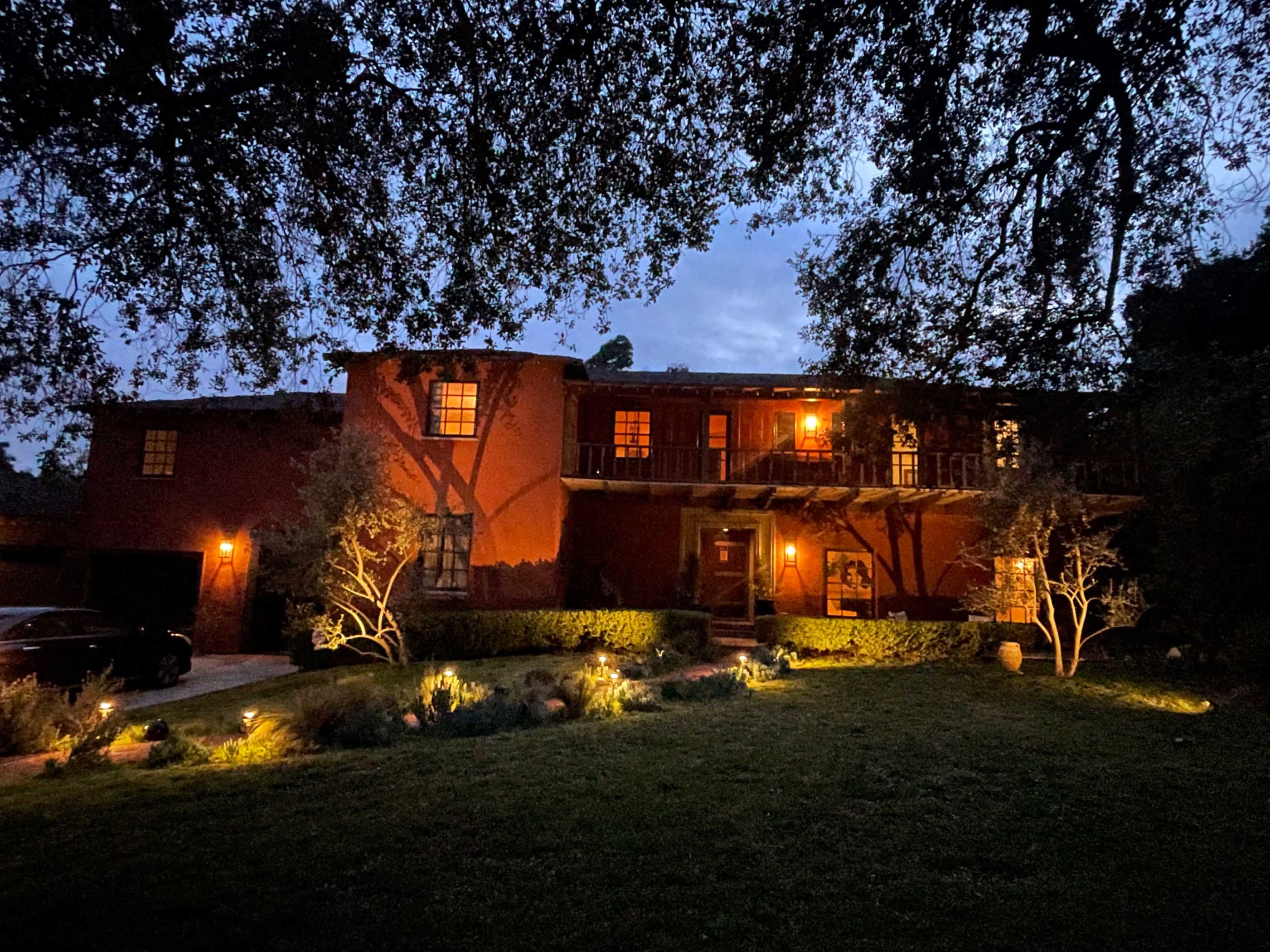 A two-story house at dusk, illuminated by outdoor lights and surrounded by green landscaping and trees.