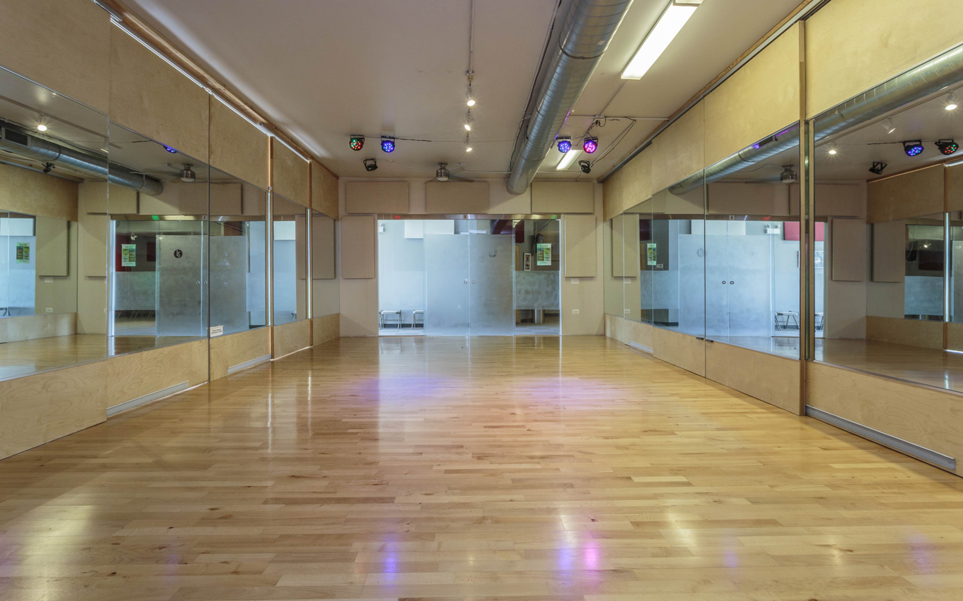 A dance studio with wooden floors and large mirrored walls, illuminated by colored lights.