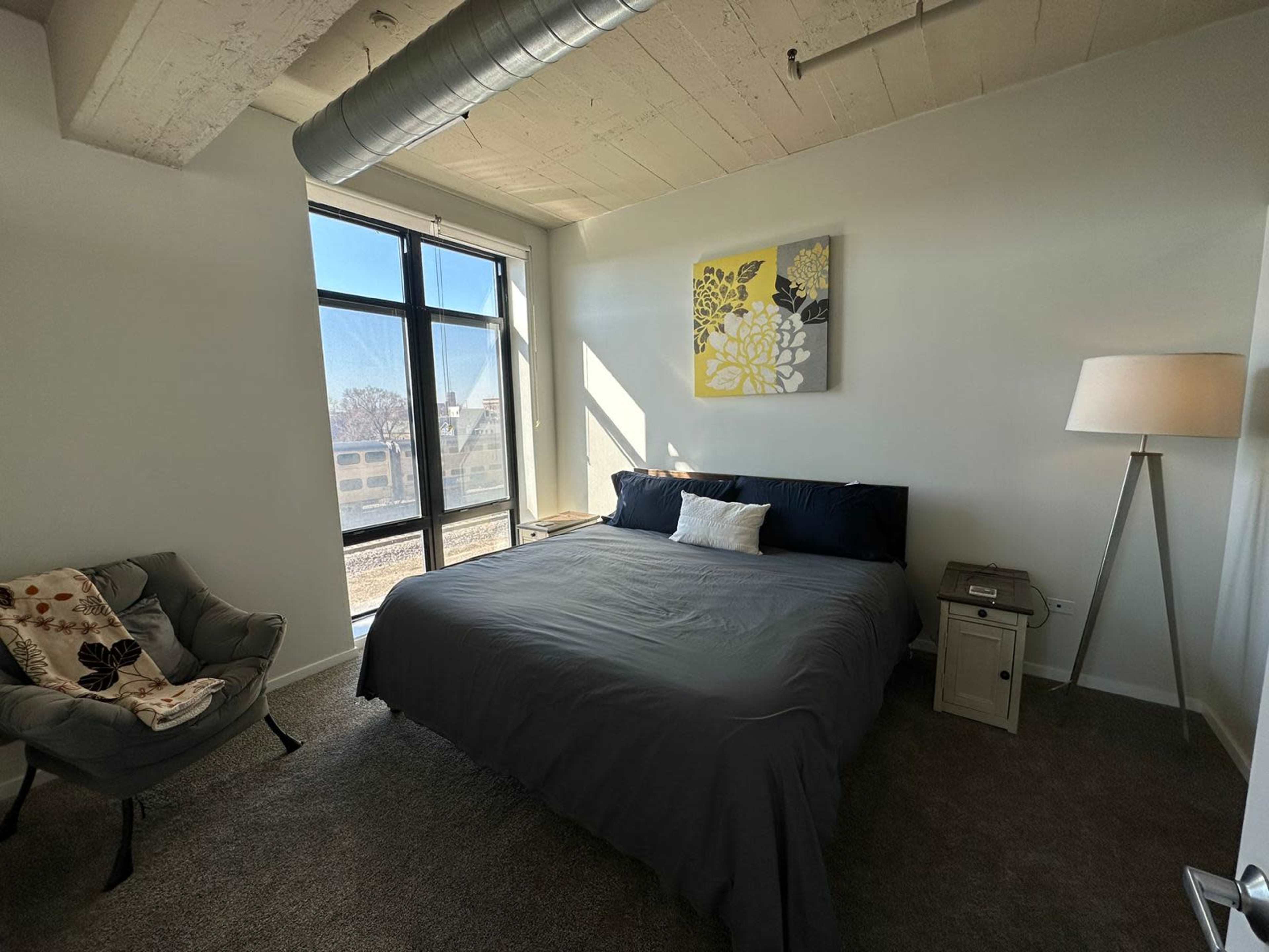A cozy bedroom featuring a large bed, a decorative wall art piece, a lamp, and a window that lets in natural light.