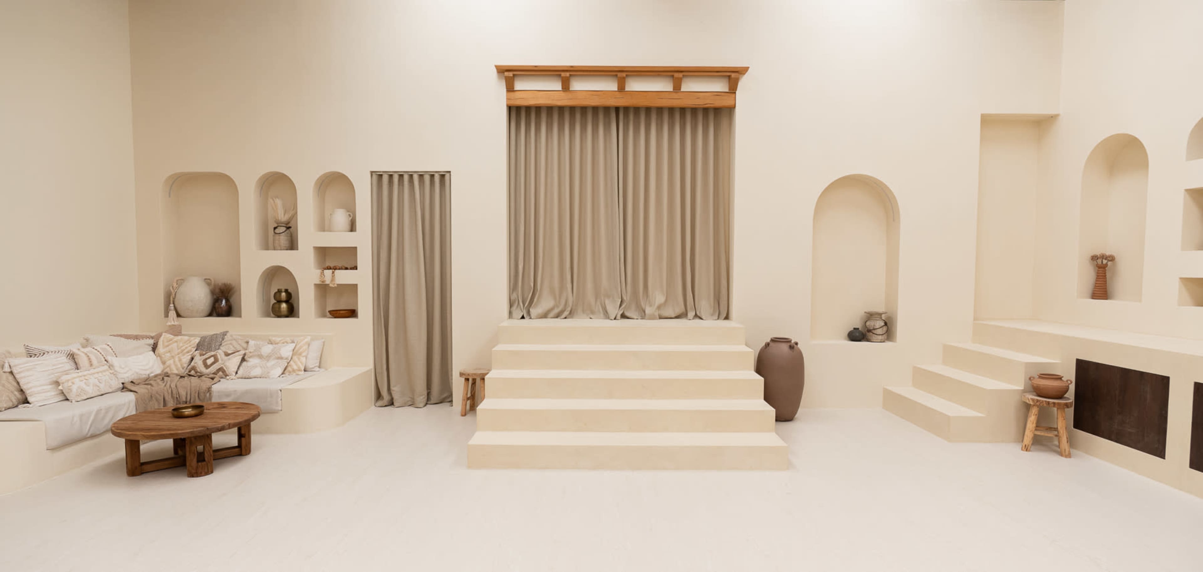 The image depicts a minimalist interior space with light beige walls, a seating area to the left, steps leading to a raised platform, and various textured niches in the walls.