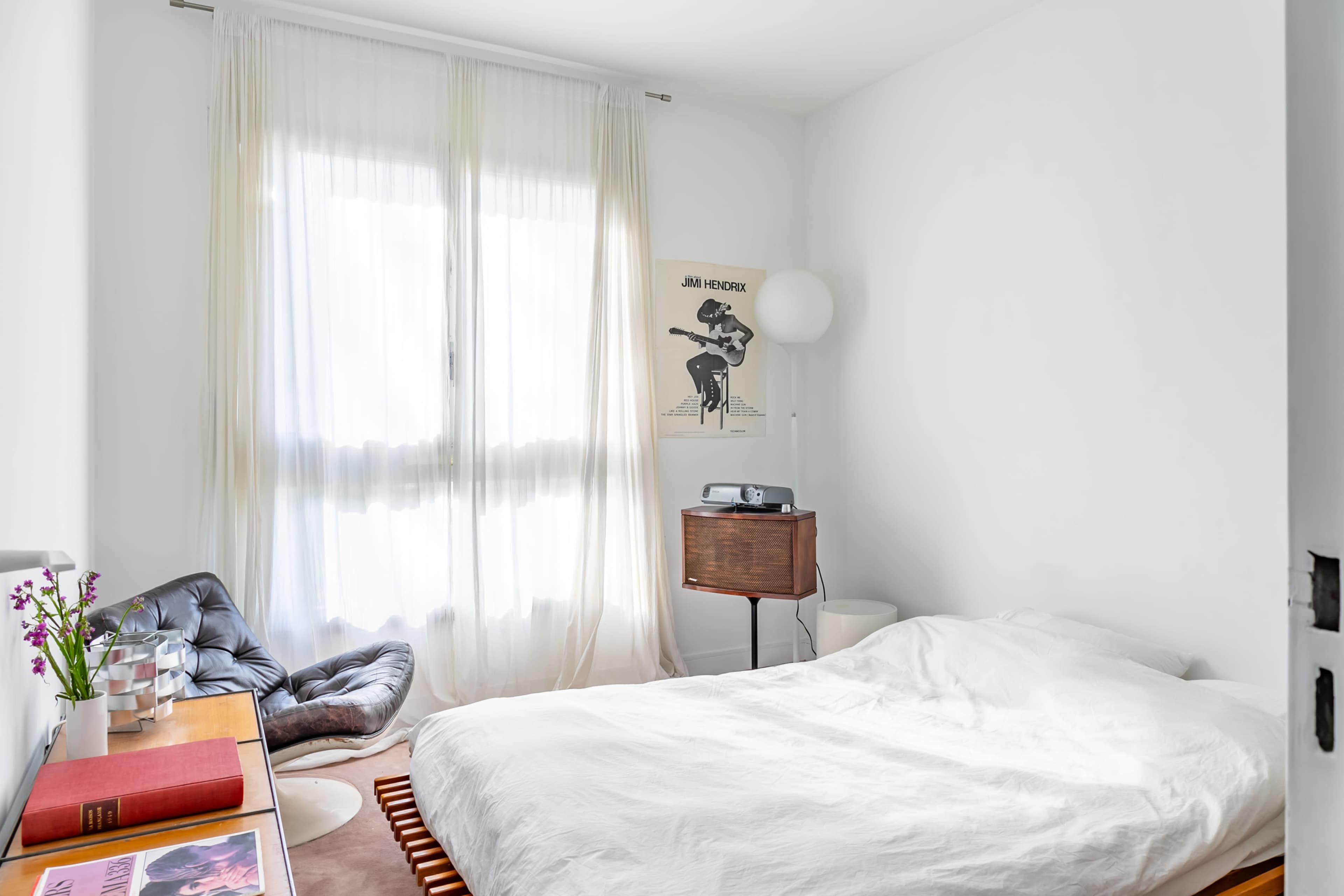 A minimalist bedroom with a white bedspread, large windows draped with sheer curtains, and a small bedside table featuring a lamp and a record player.