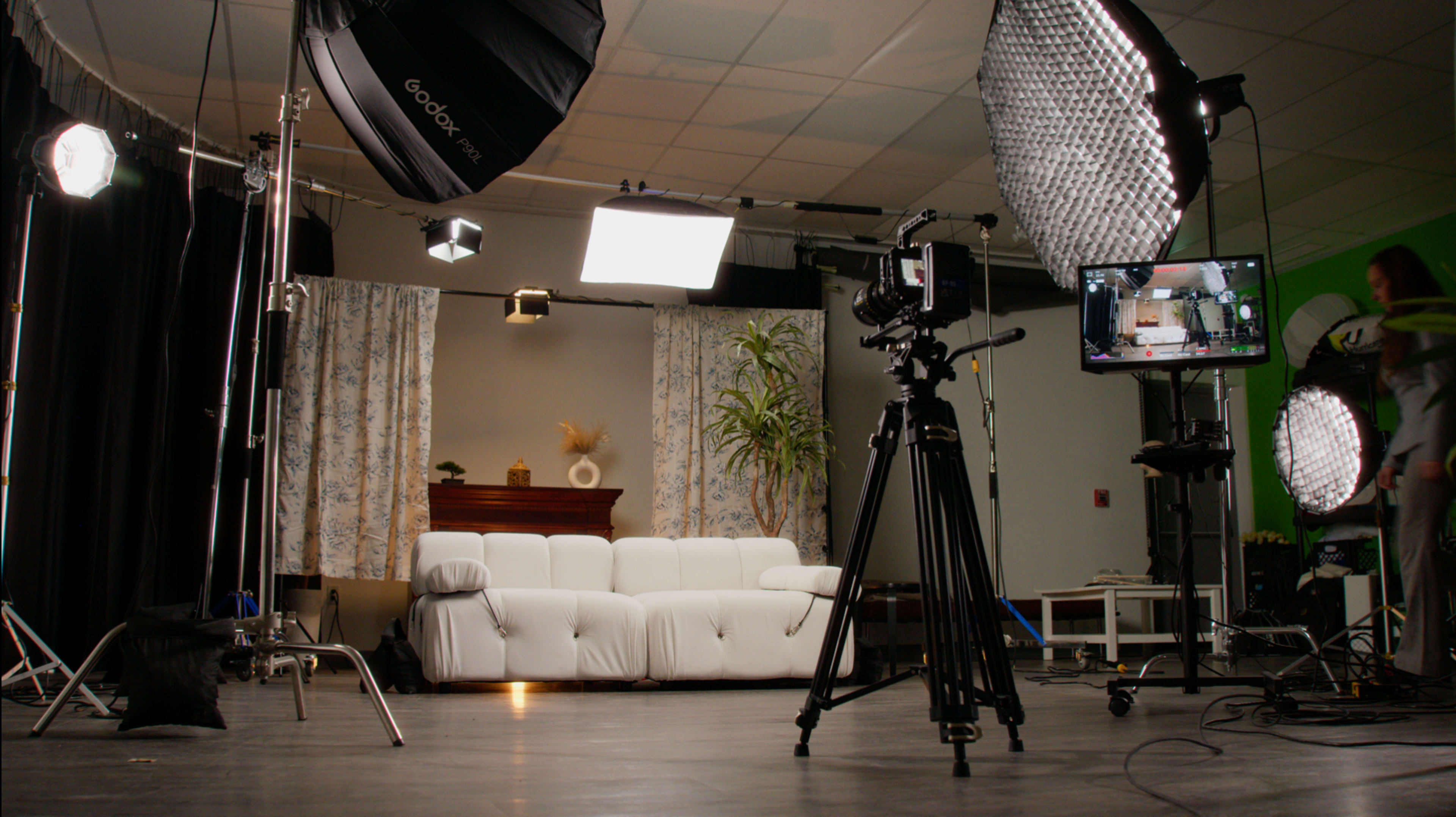 The image depicts a well-equipped studio set featuring a white couch, curtains, and various lighting and camera equipment arranged around the space.