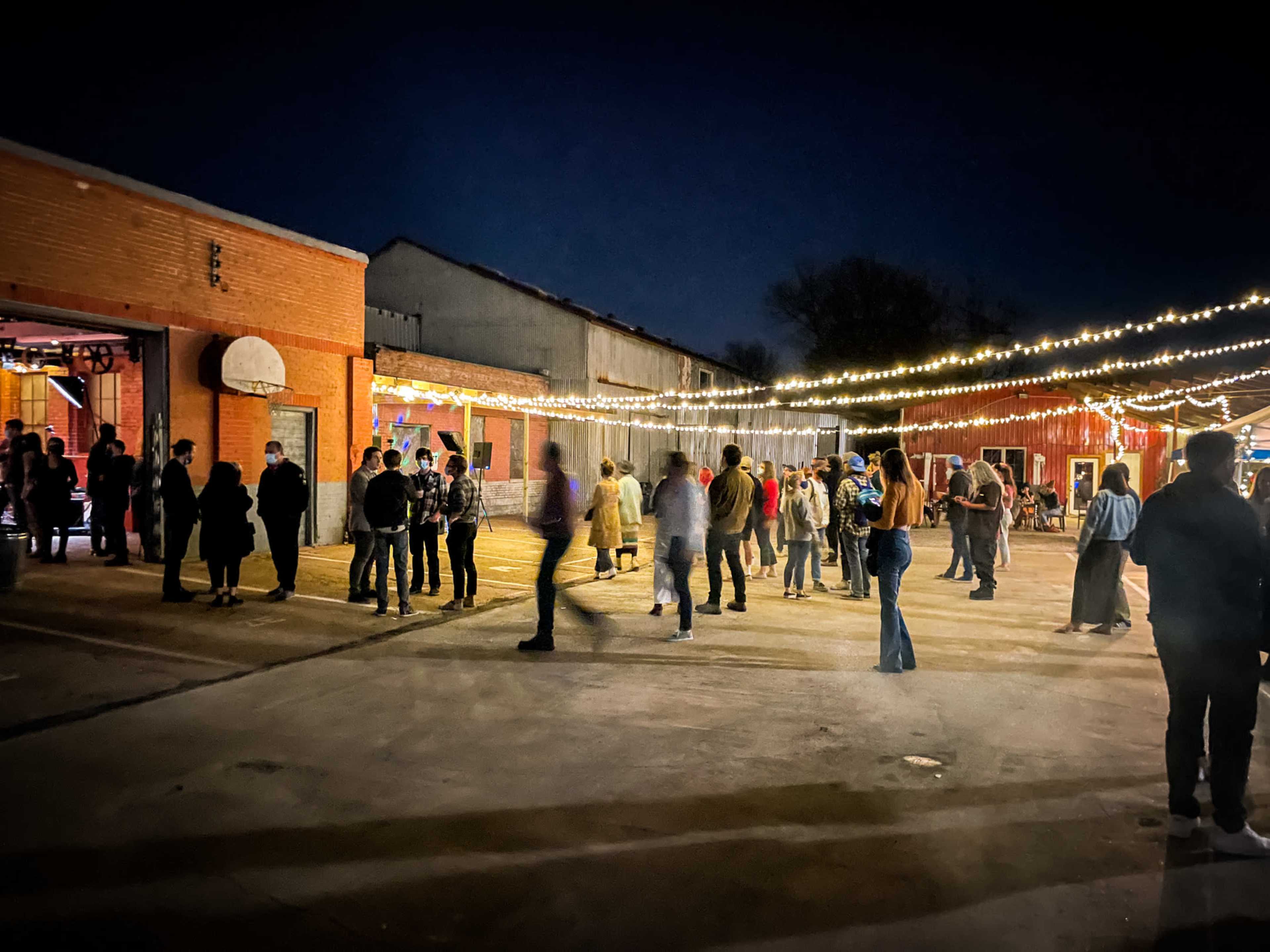 A crowd of people stands in a lit outdoor area with string lights above, surrounded by brick buildings at night.