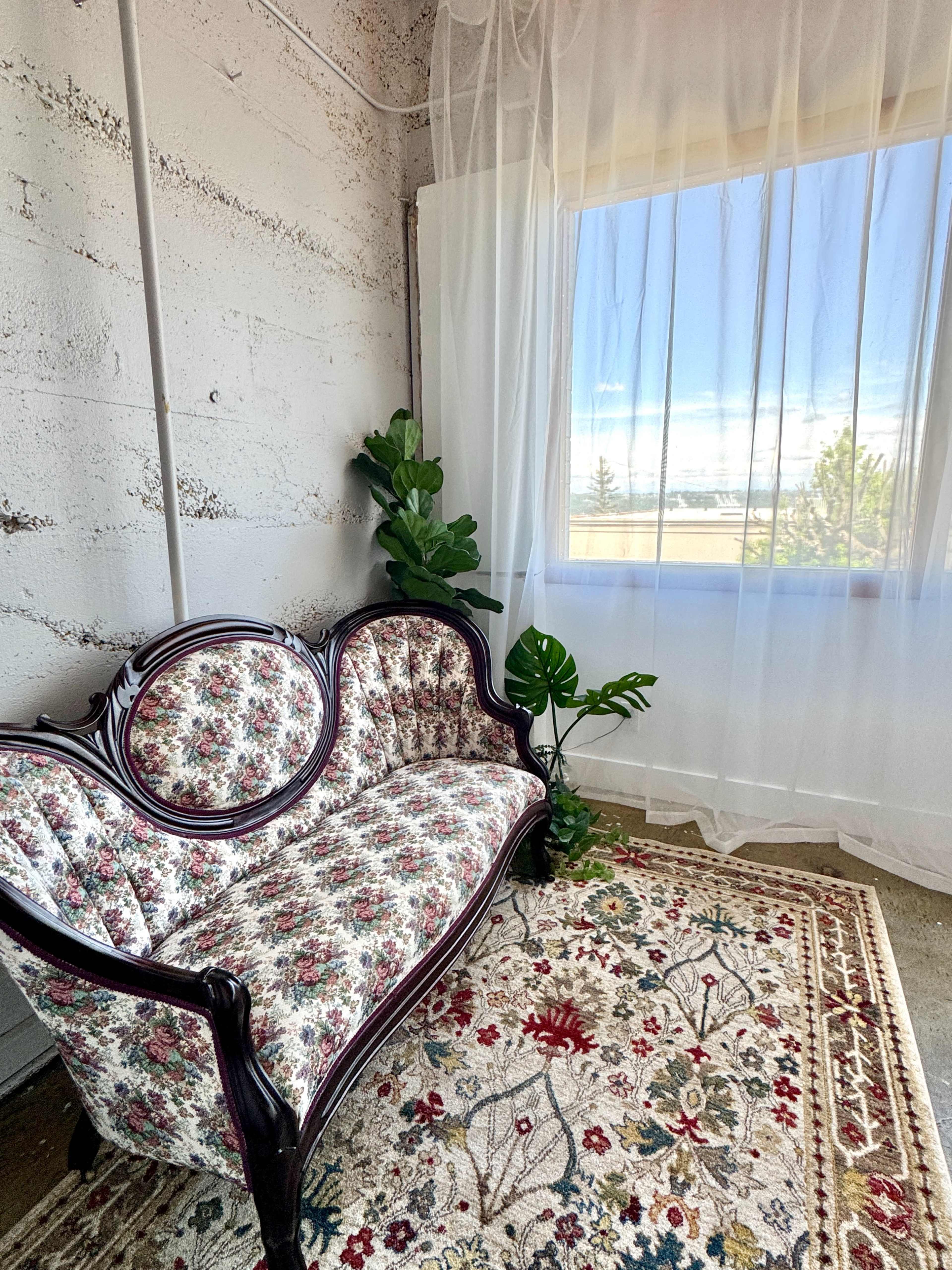 The image shows a vintage floral-patterned loveseat placed near a window with sheer curtains, next to a potted plant and a decorative rug on the floor.