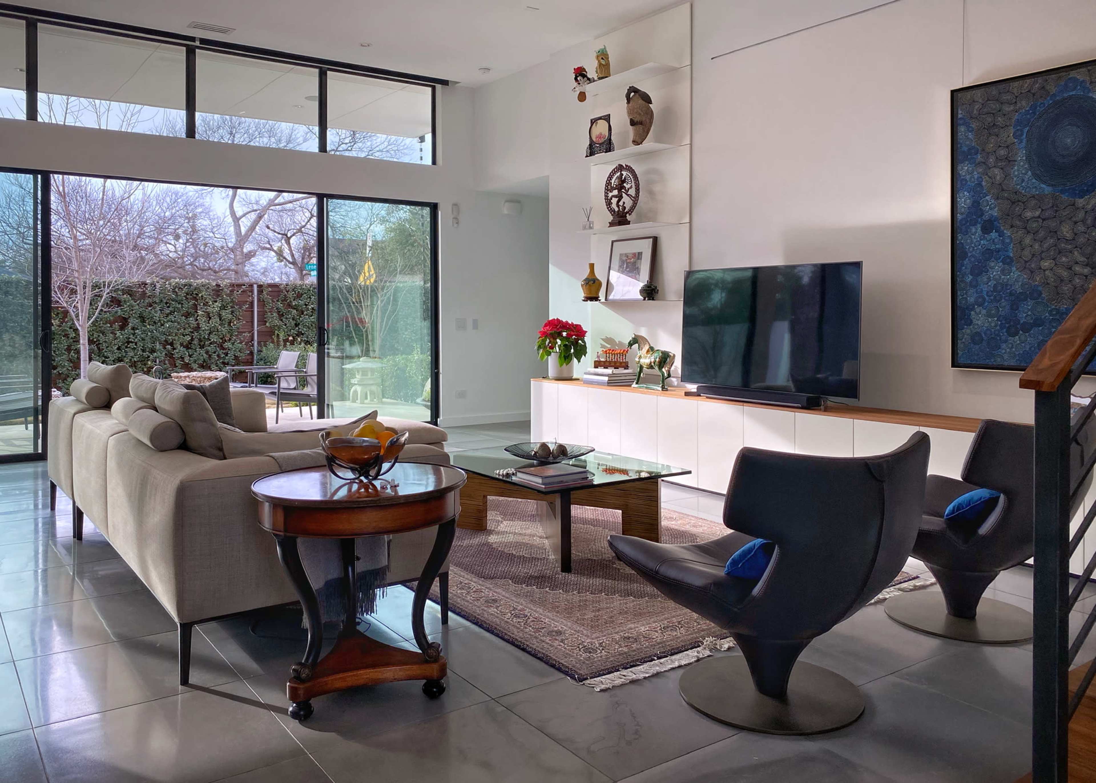 A modern living room features a sofa, two black chairs, a glass coffee table, and a television on a white cabinet, with large windows offering a view of the garden outside.