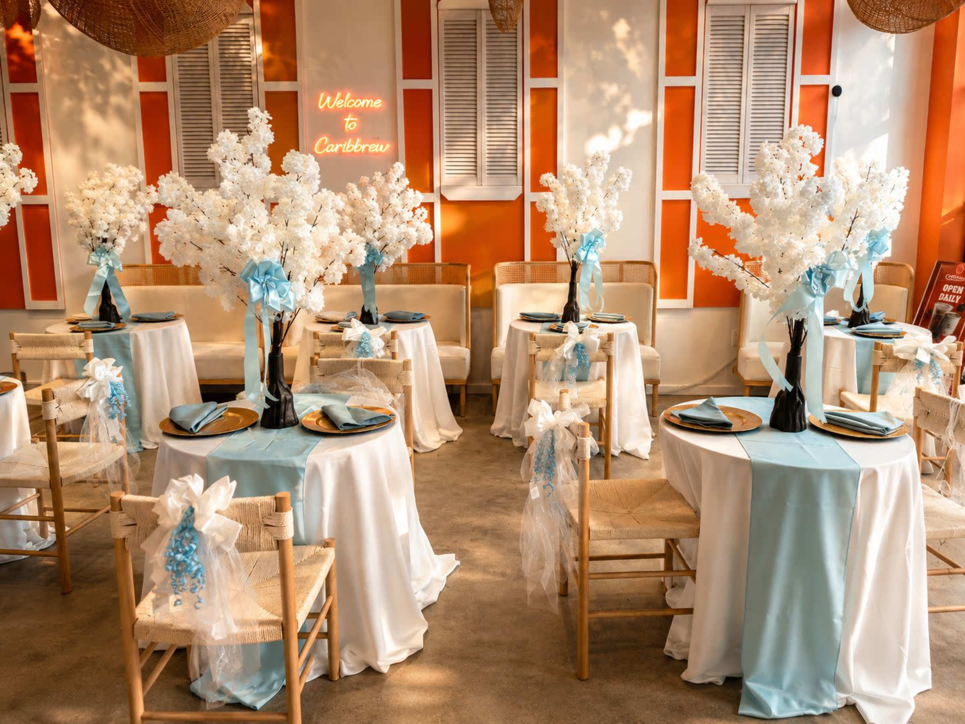 The image shows an elegantly arranged dining area with tables adorned with light blue and white decorations, featuring tall floral centerpieces and neatly set dinnerware.