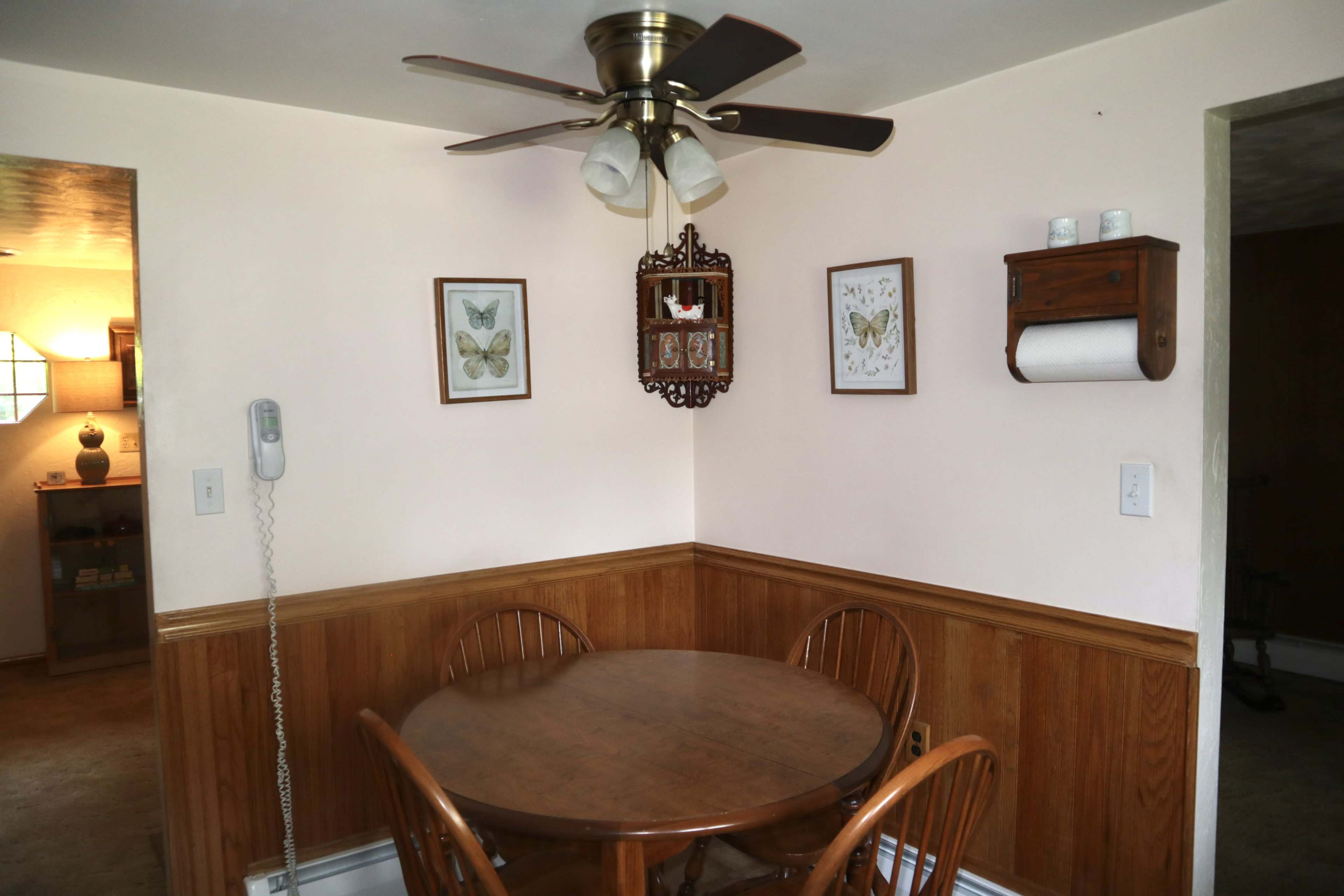 The image shows a small dining area with a wooden table and chairs, a ceiling fan, and framed art on the walls.