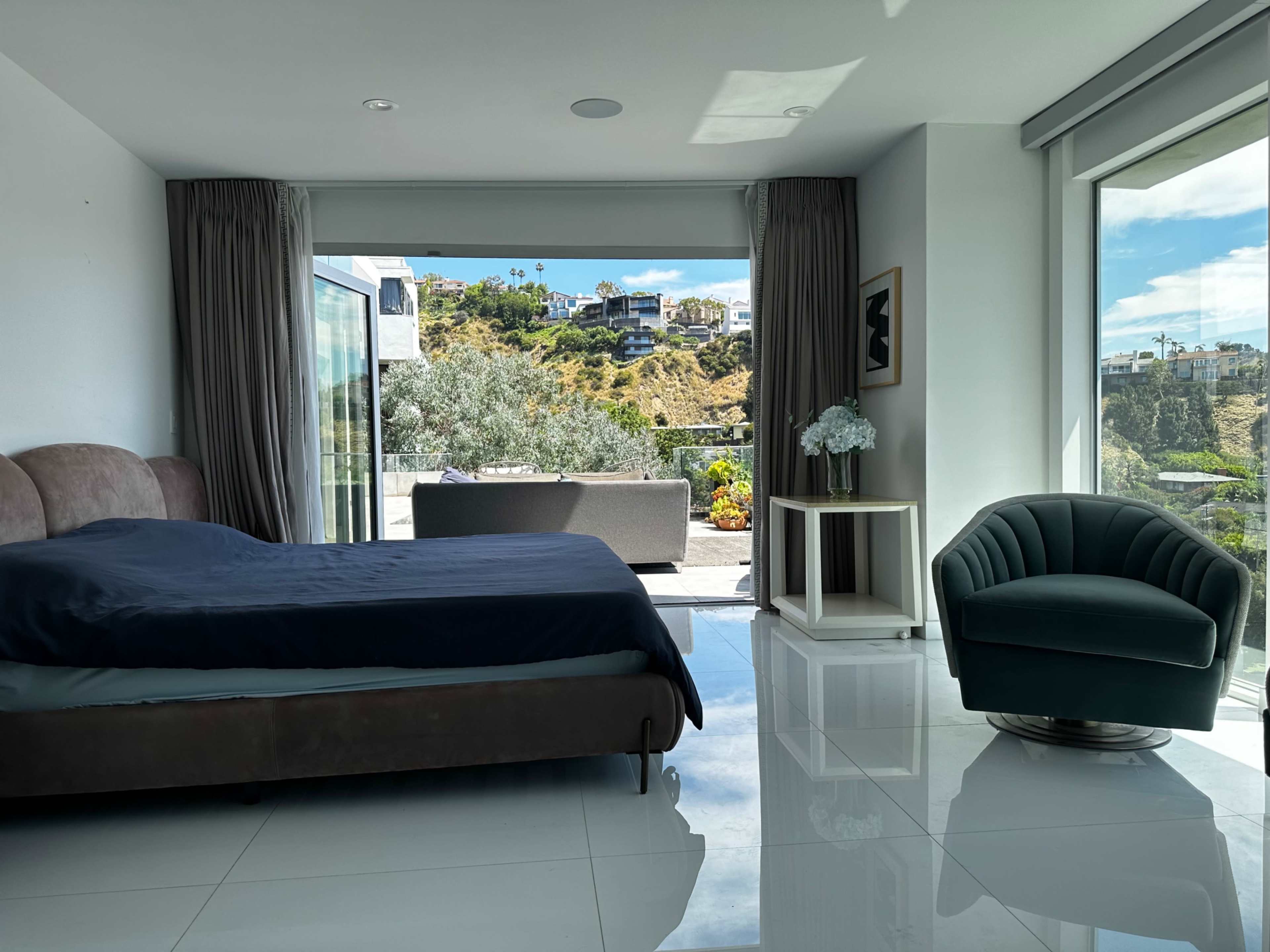 A modern bedroom features a bed and a green chair, with large windows showcasing a hillside view.