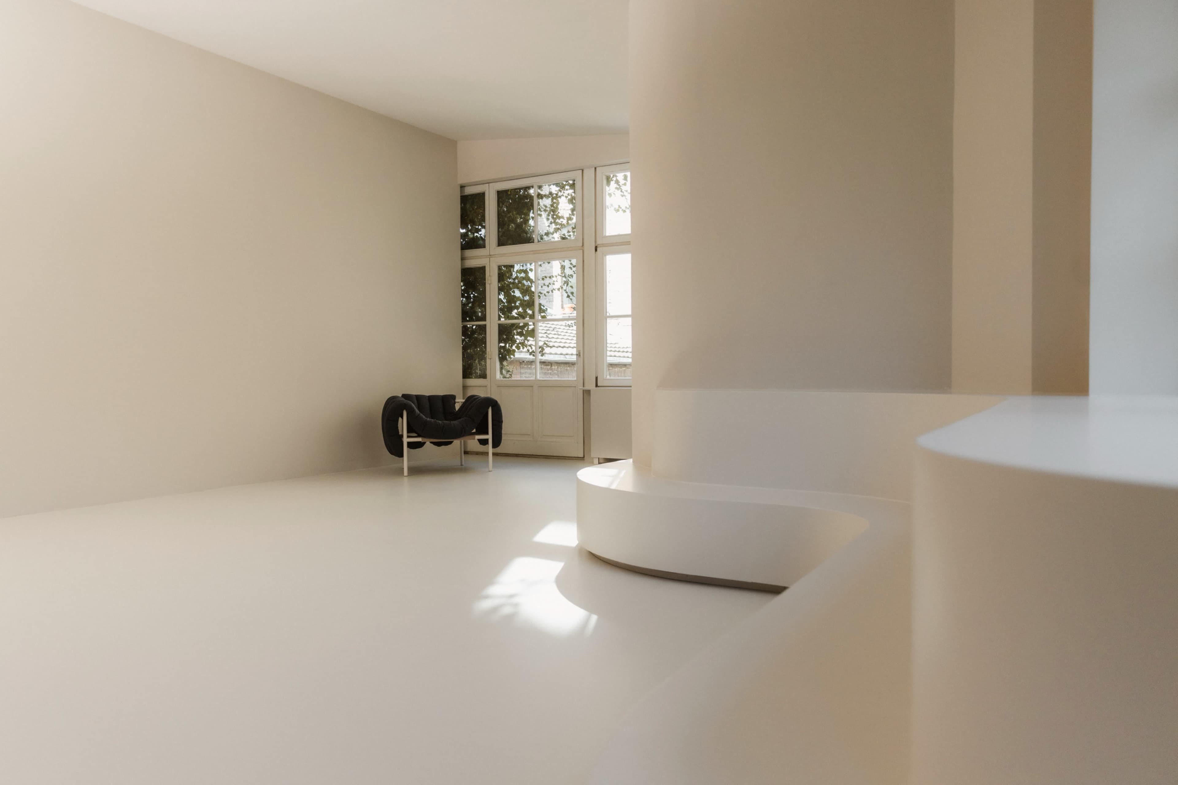The image shows a minimalist interior space with light-colored walls, a curved white feature, and a modern black chair near a window.