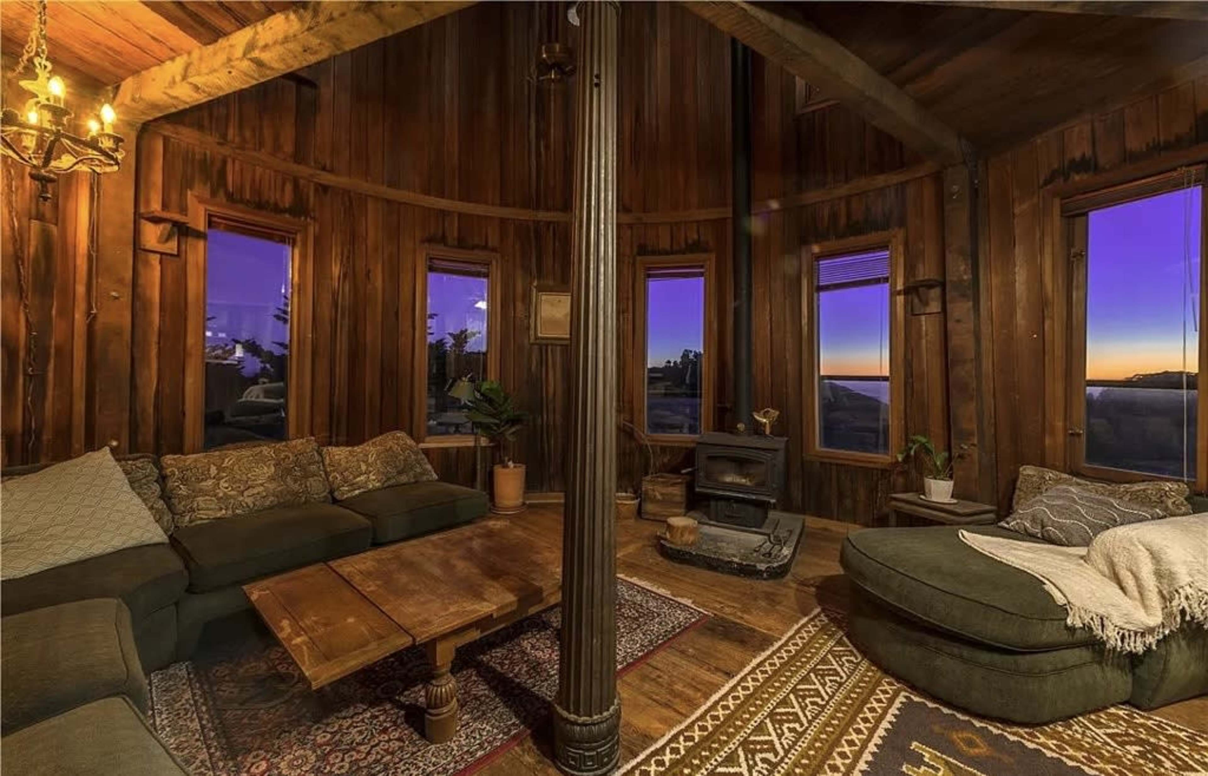 The interior of a rustic living room features wooden walls, a central pole, a wood stove, and large windows showcasing a twilight view.
