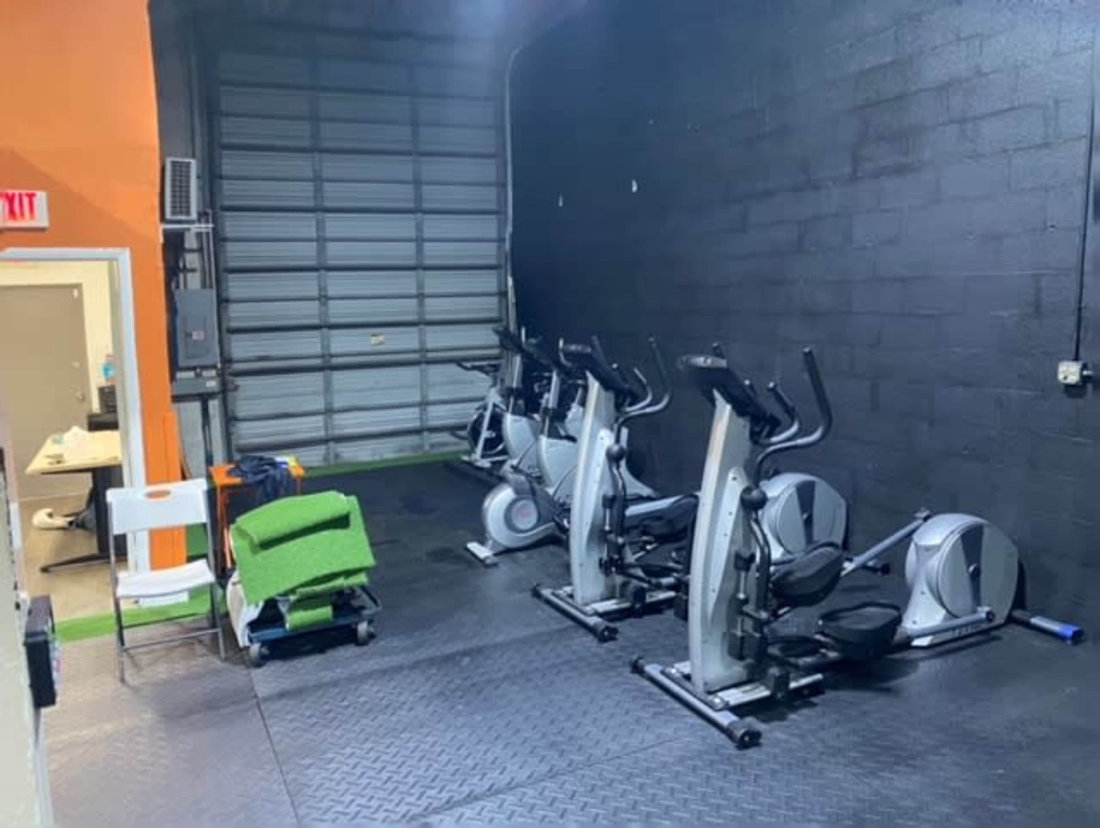A small gym area with several exercise machines, including ellipticals, on a rubber mat near a garage door.