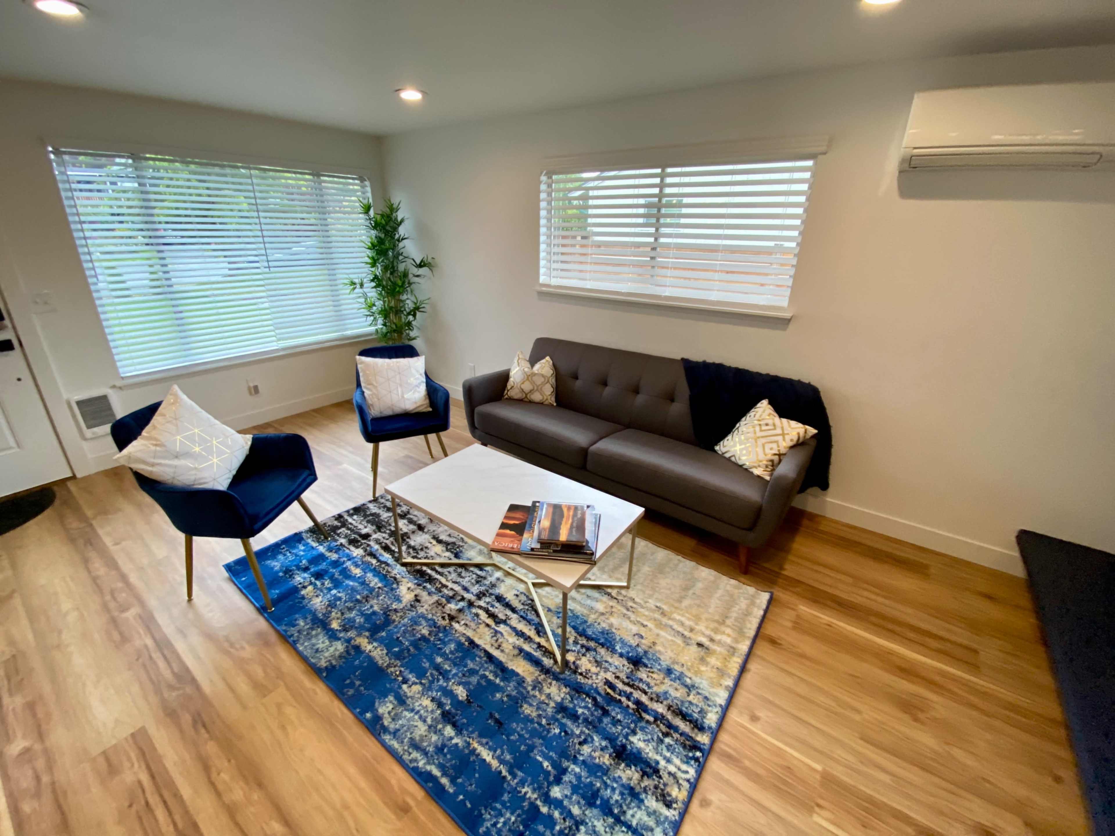 A modern living room featuring a gray sofa, two dark blue chairs, a coffee table with a magazine, and a large rug, all illuminated by natural light from the windows.