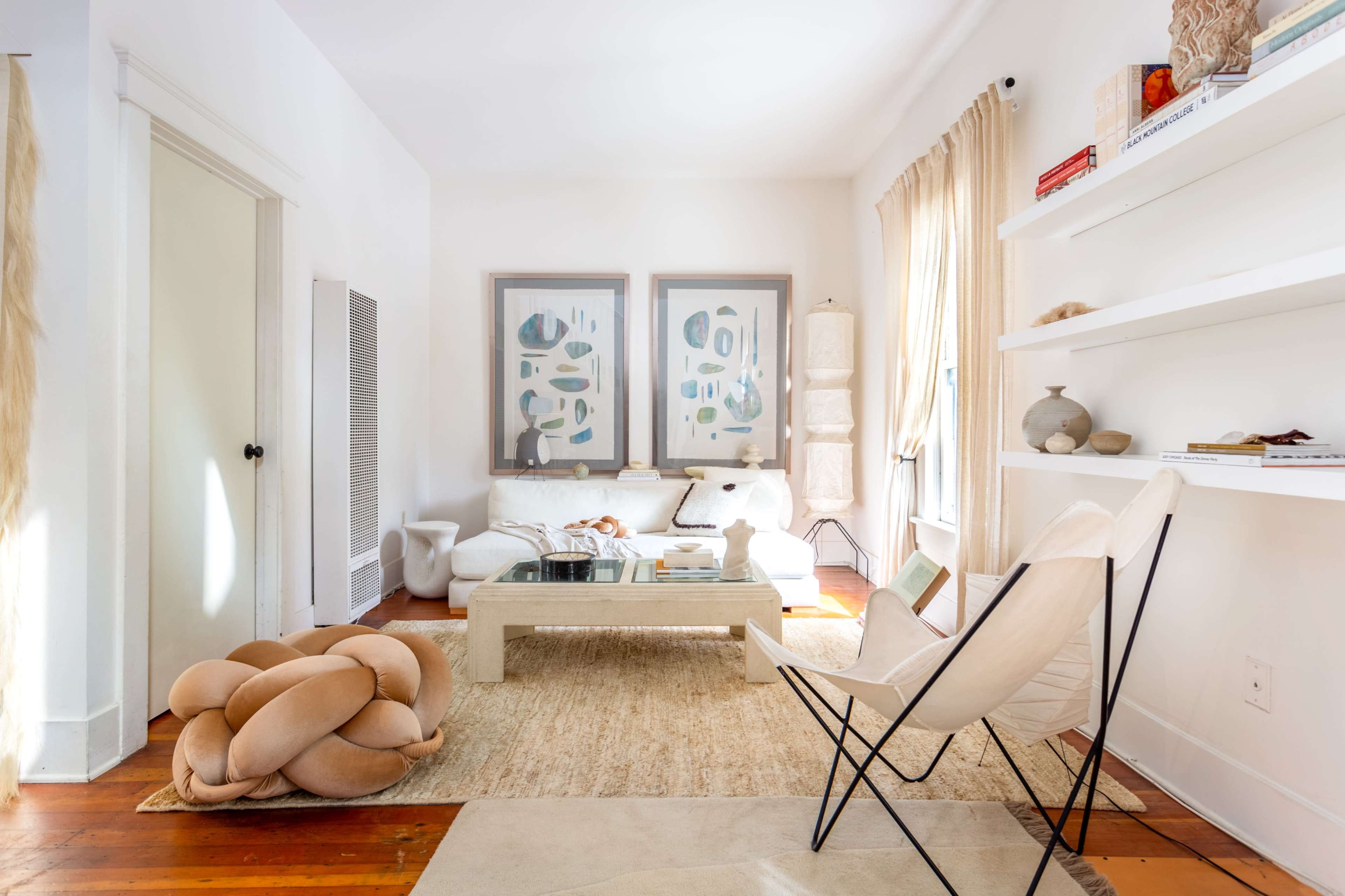 A bright living room features a minimalist design with a glass coffee table, a large plush pouf, and two framed artworks on the wall.