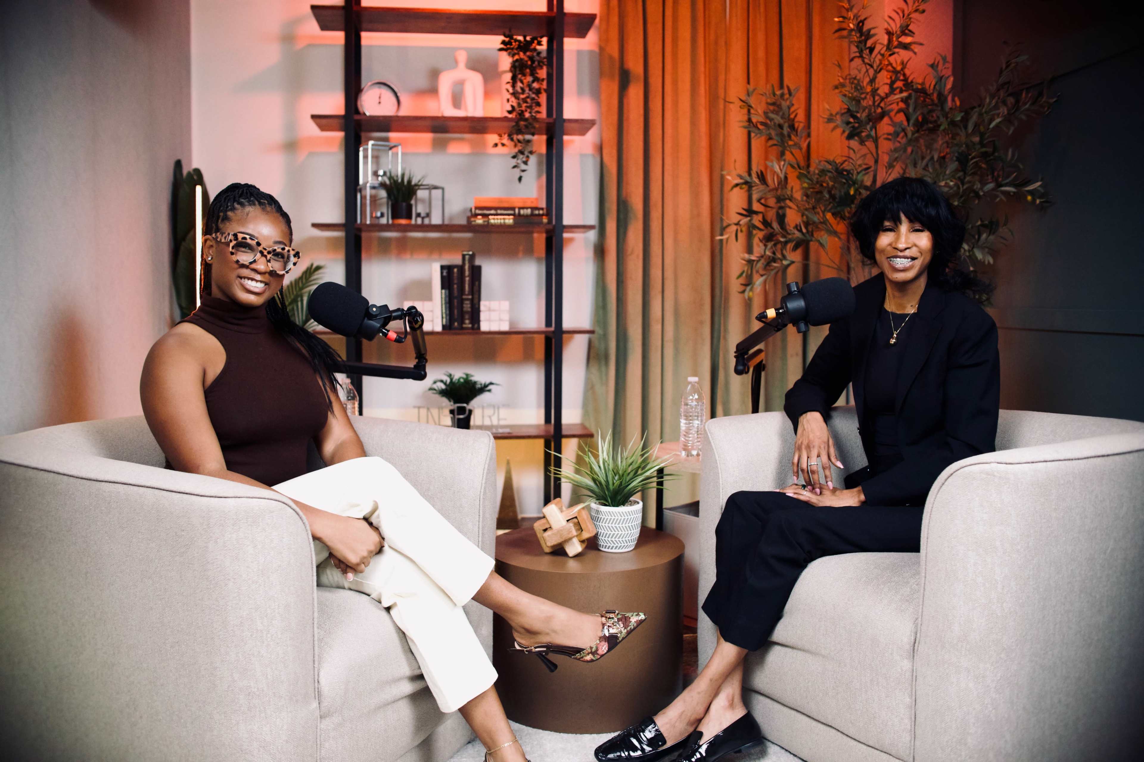 Two women sit in armchairs with microphones in a cozy podcast studio, surrounded by plants and decor.