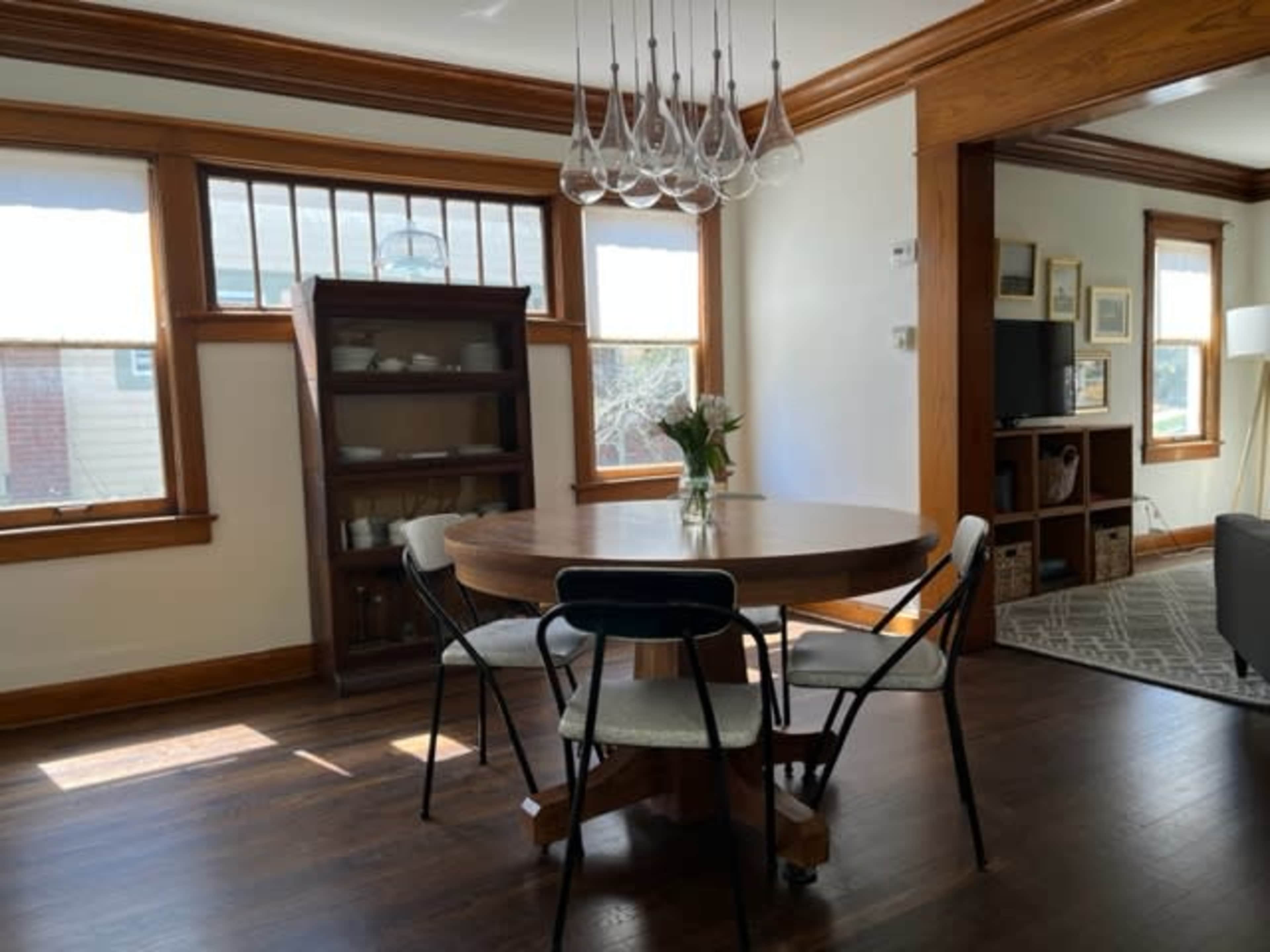 The image shows a dining area featuring a round wooden table surrounded by chairs, with a vase of flowers on the table and a light fixture overhead.