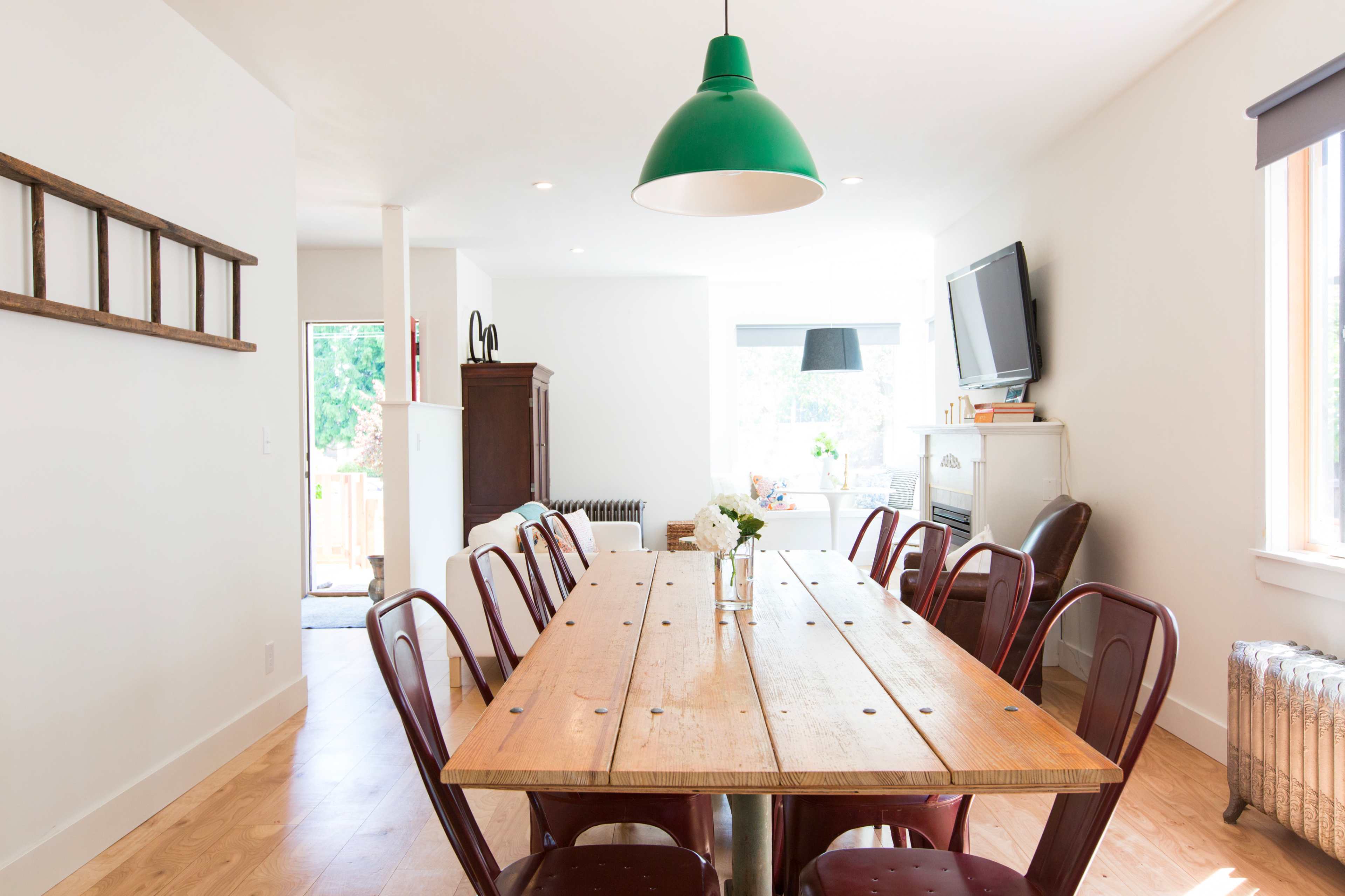 A long wooden dining table with metal chairs is set in a bright, minimalistic room featuring a green pendant light and a large window.
