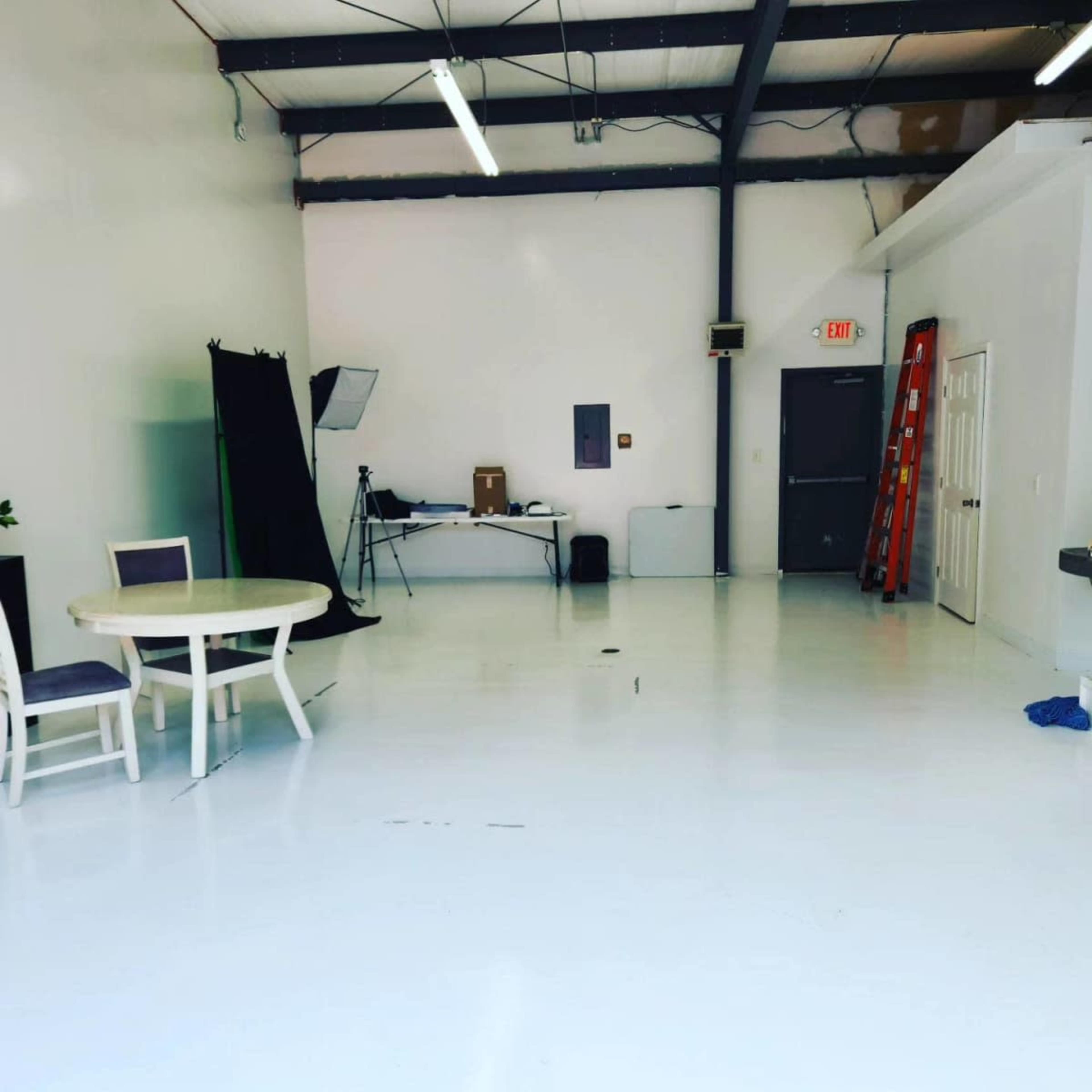 The image shows a spacious, empty room with white walls and a polished floor, featuring a round table with chairs, a ladder, and various pieces of equipment in the background.