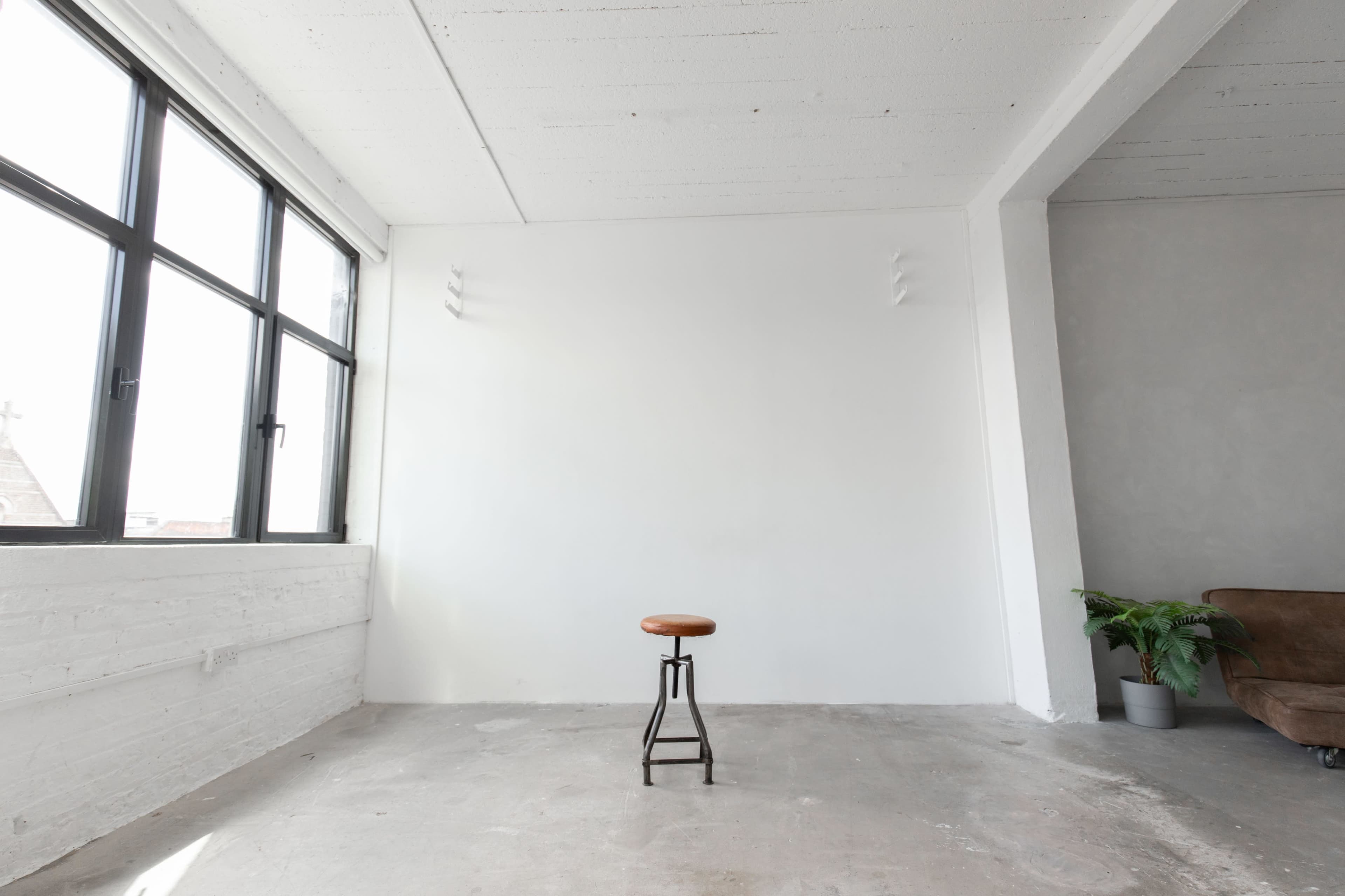 A minimalist interior space with a single wooden stool positioned in front of a blank white wall, large windows, and a potted plant in the corner.