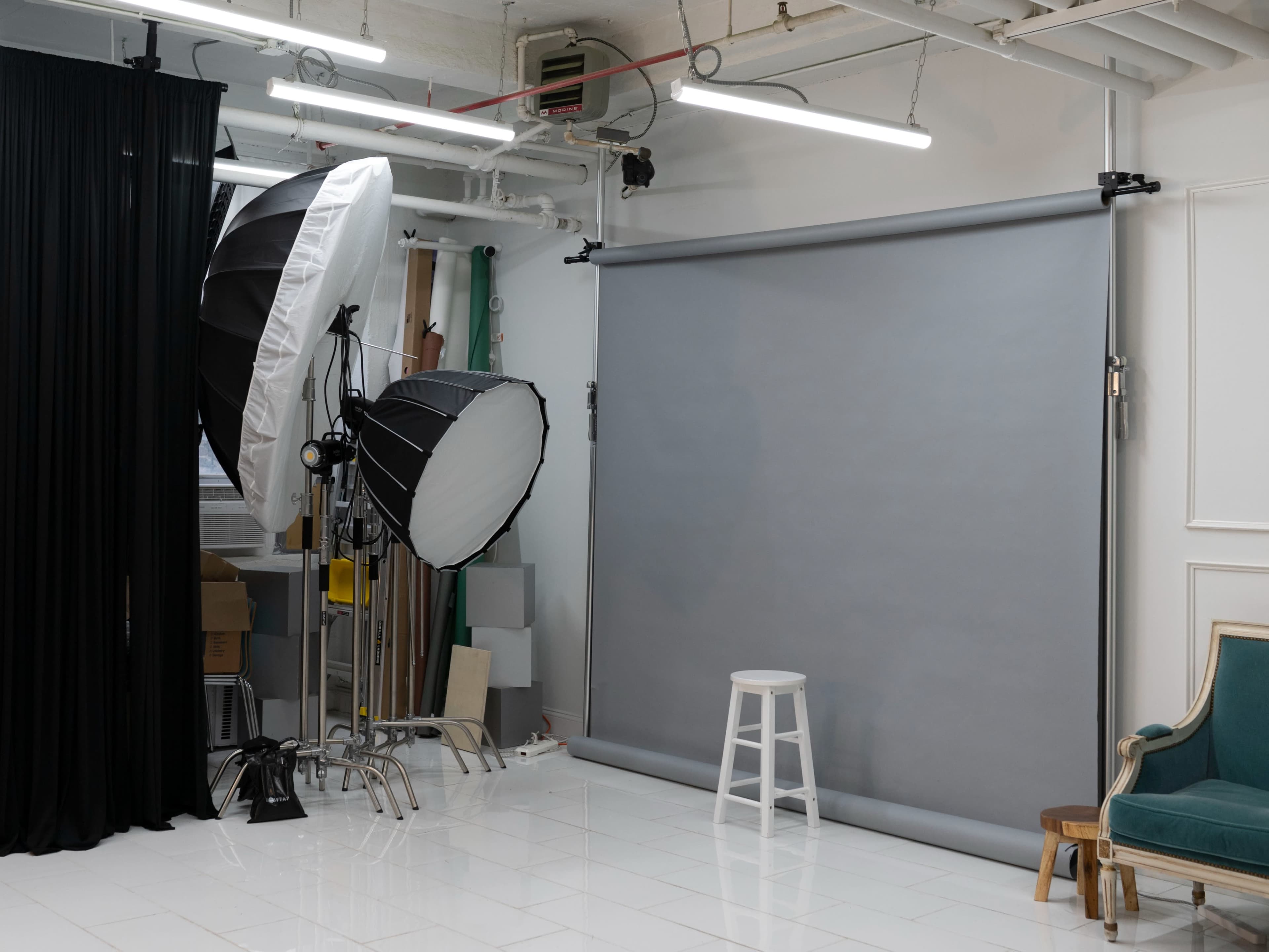 The image shows a photography studio featuring a gray backdrop, softbox lighting equipment, and a white stool.