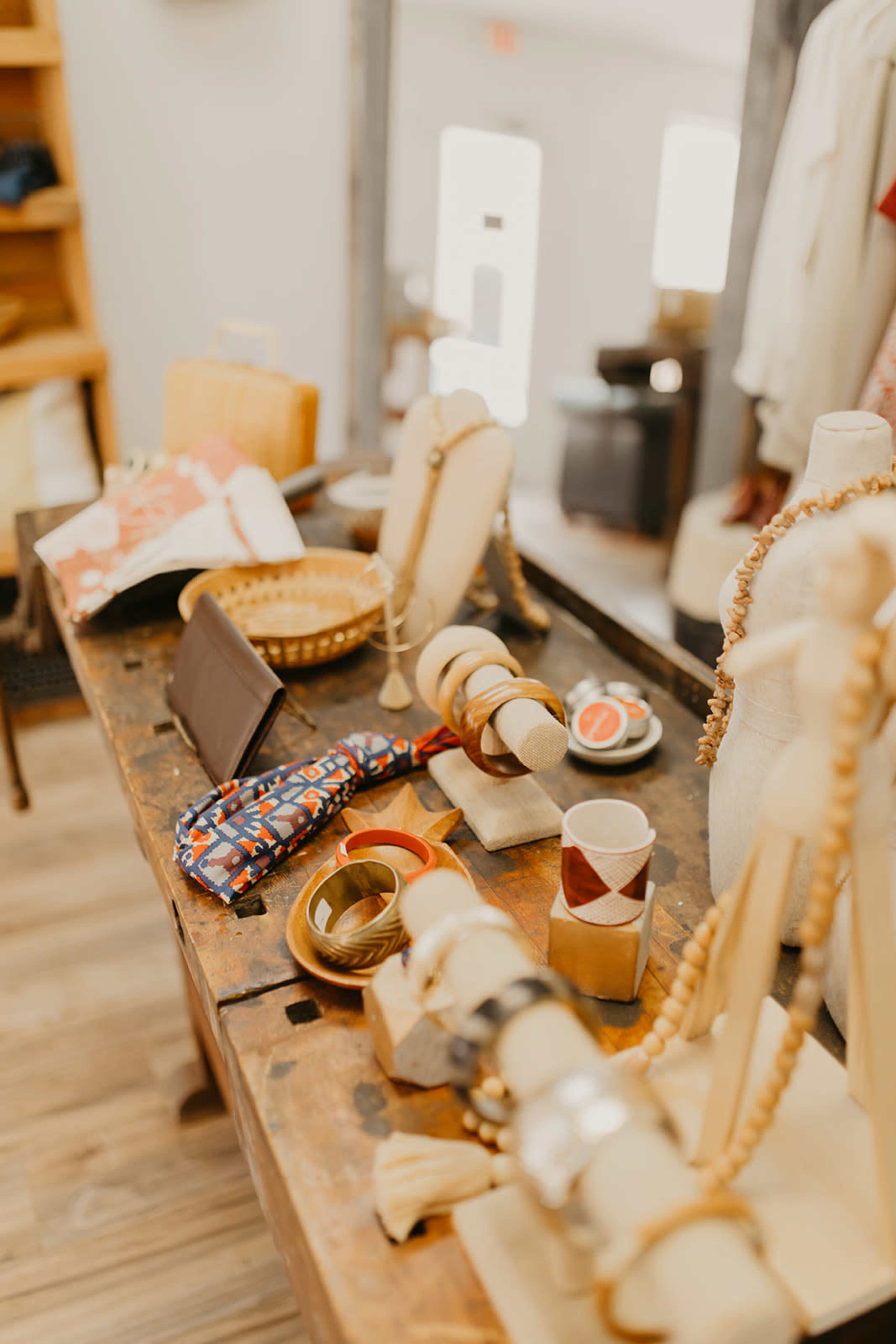 A wooden table displays various handmade accessories, including bracelets, fabric items, and a basket, in a boutique setting.