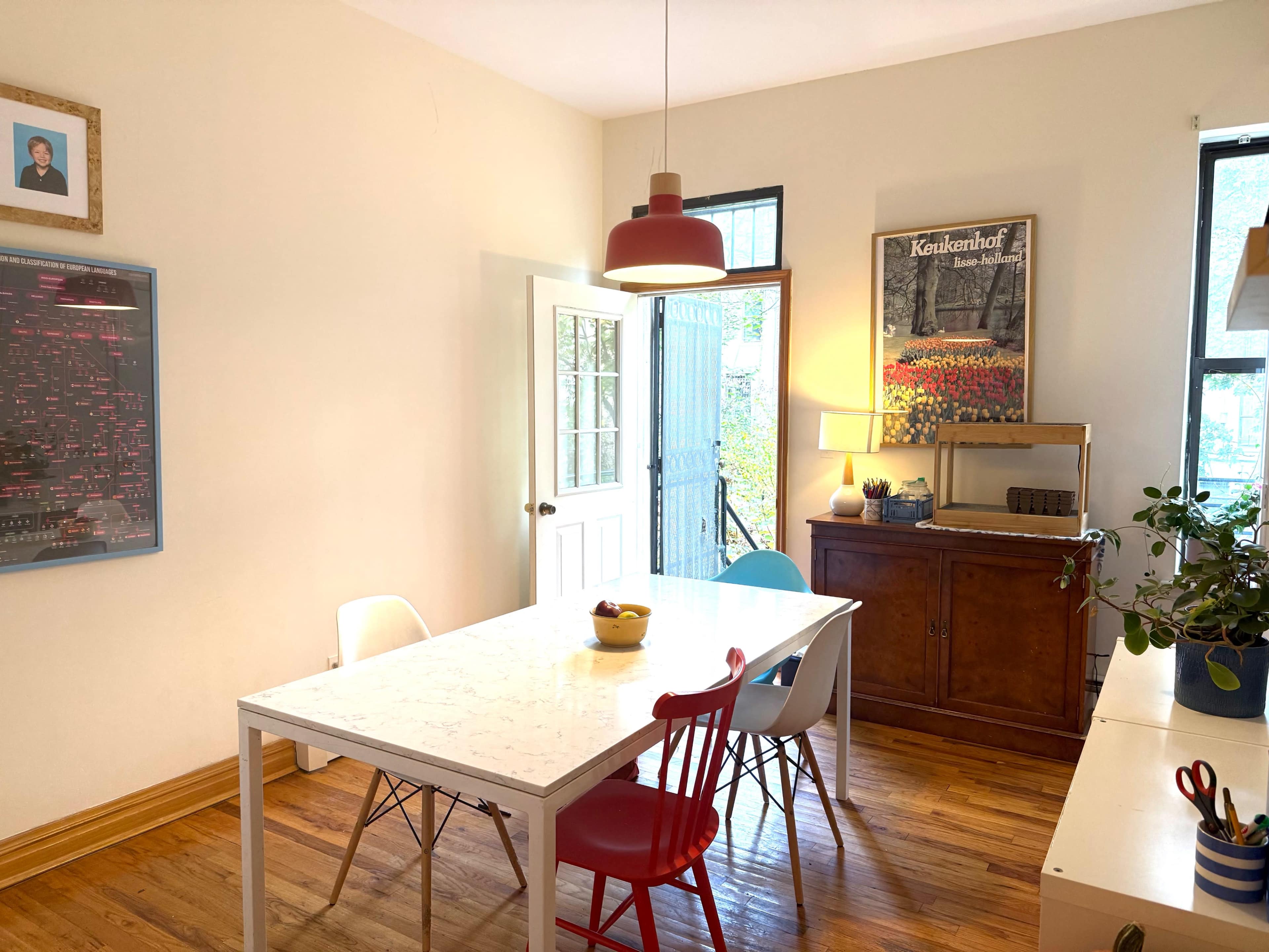 A bright dining area features a white table surrounded by colorful chairs, with a wooden sideboard against the wall and a door leading outside.