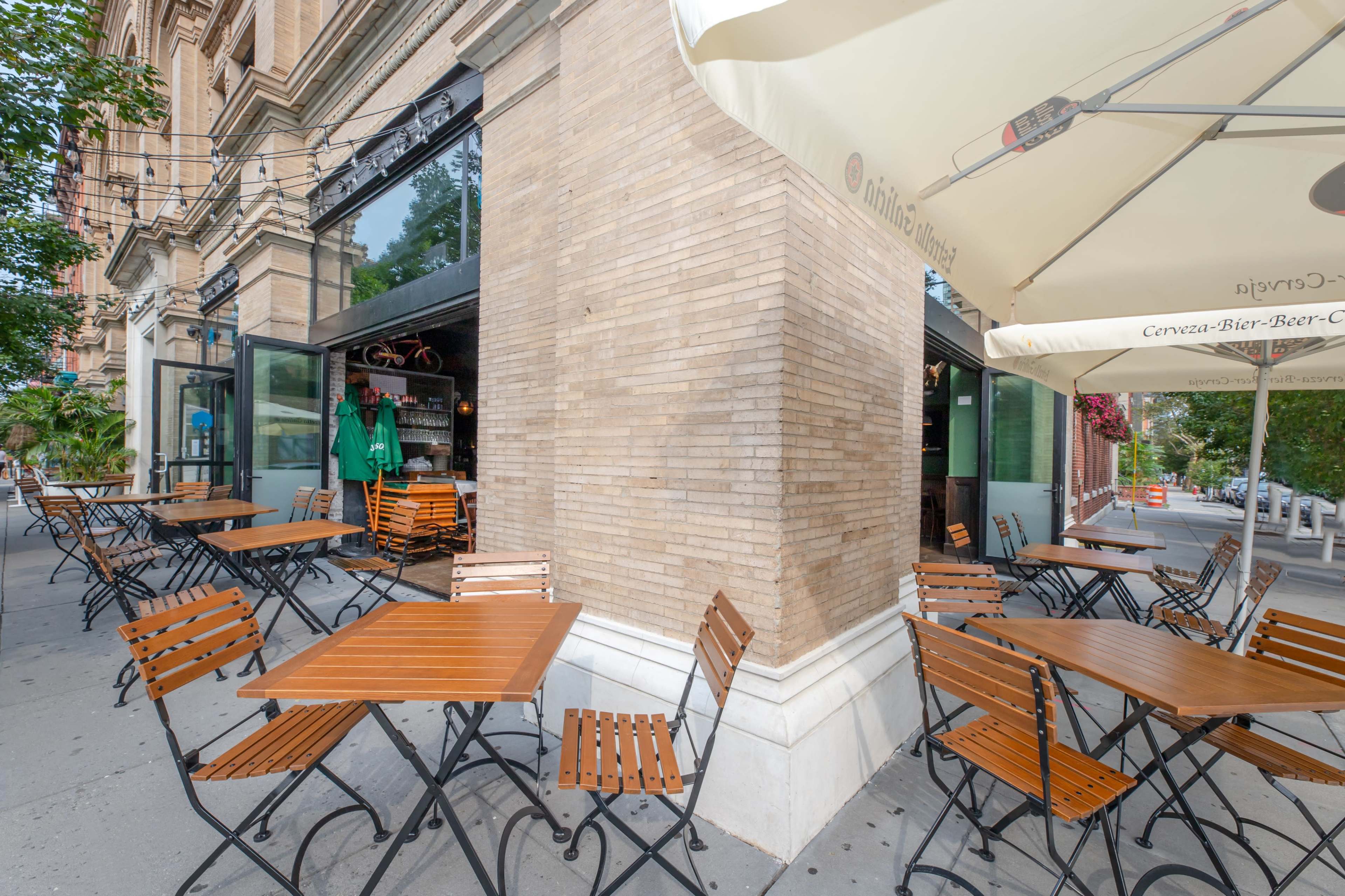 A cafe with wooden tables and chairs is located outside on a sidewalk next to a brick building.
