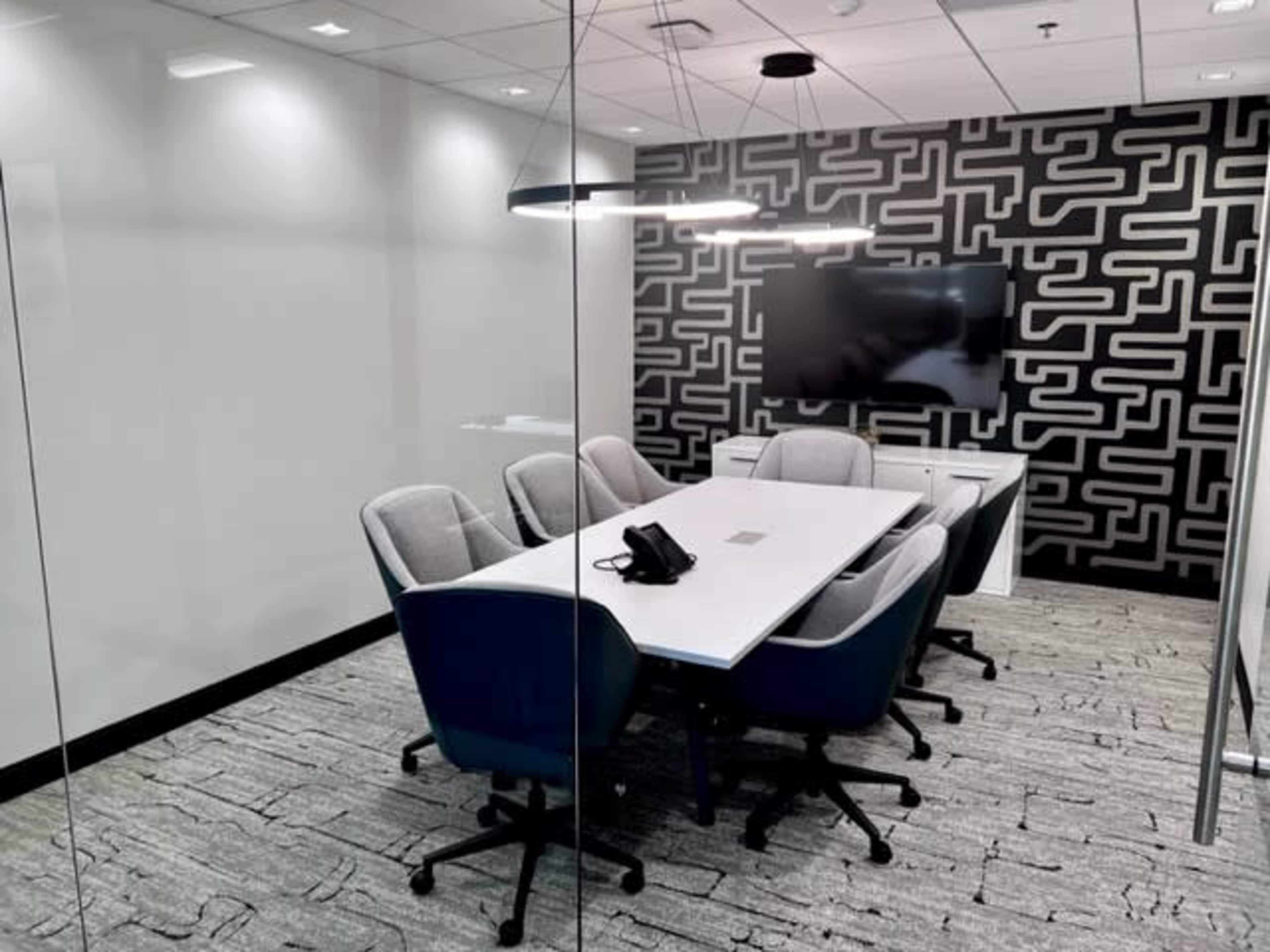A modern conference room with a large table, six chairs, a television on the wall, and a glass partition.