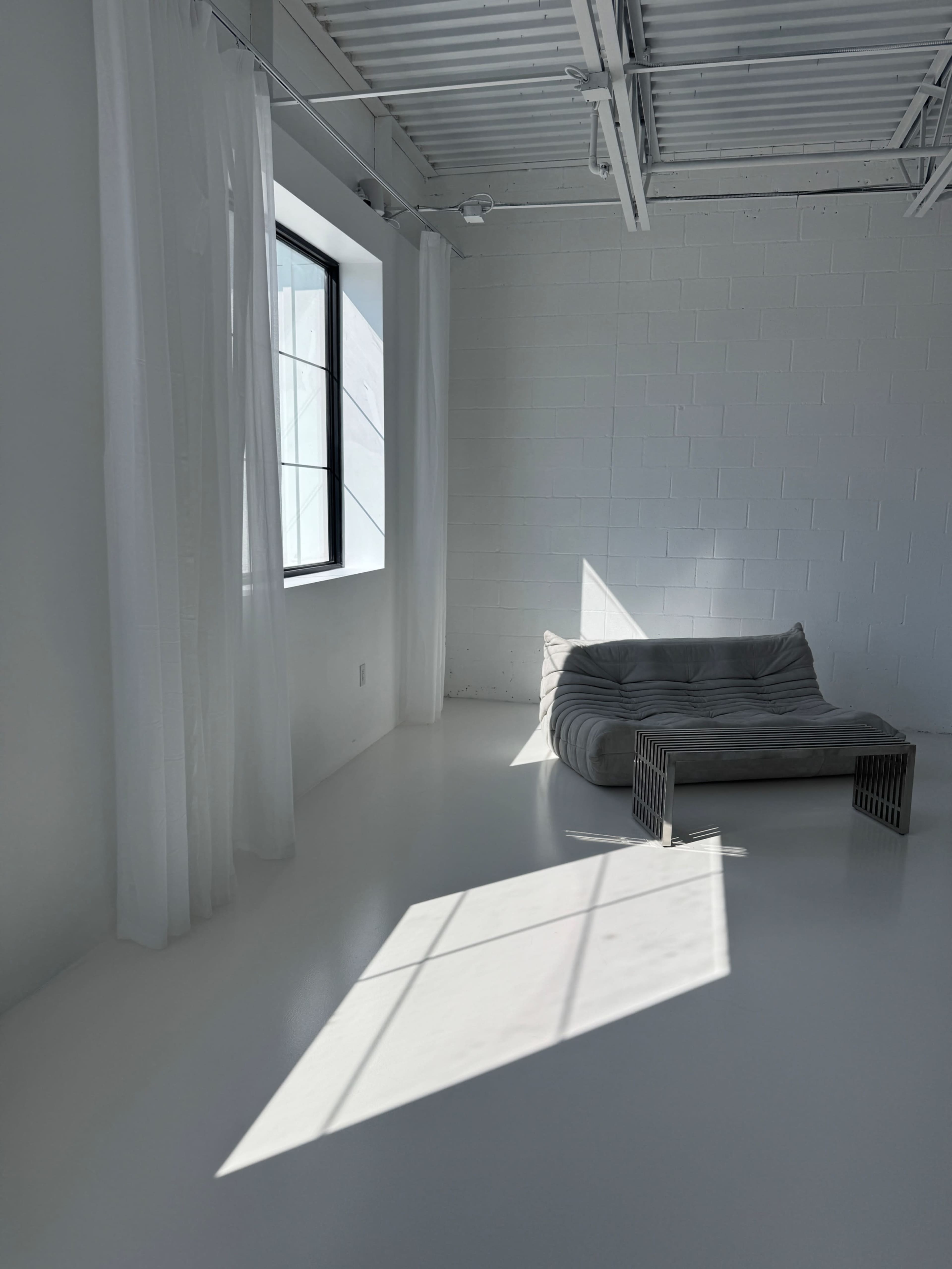 The image shows a minimalist room with a gray sofa, sheer white curtains, and a large window casting a shadow on the polished white floor.